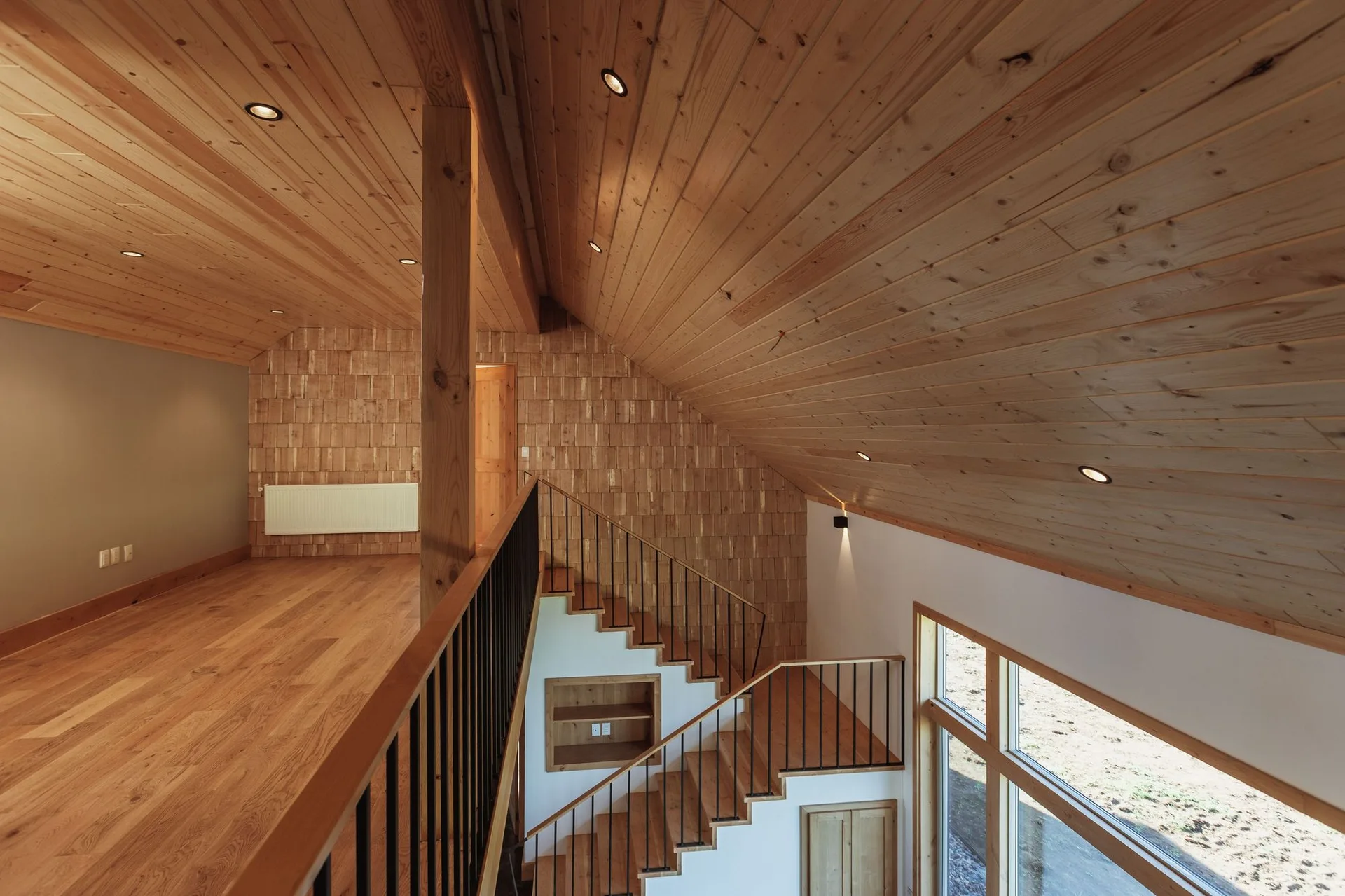 An open loft mezzanine features light wood plank floors, a sloped wooden ceiling, a shingle-clad accent wall, and a staircase with black metal railings.