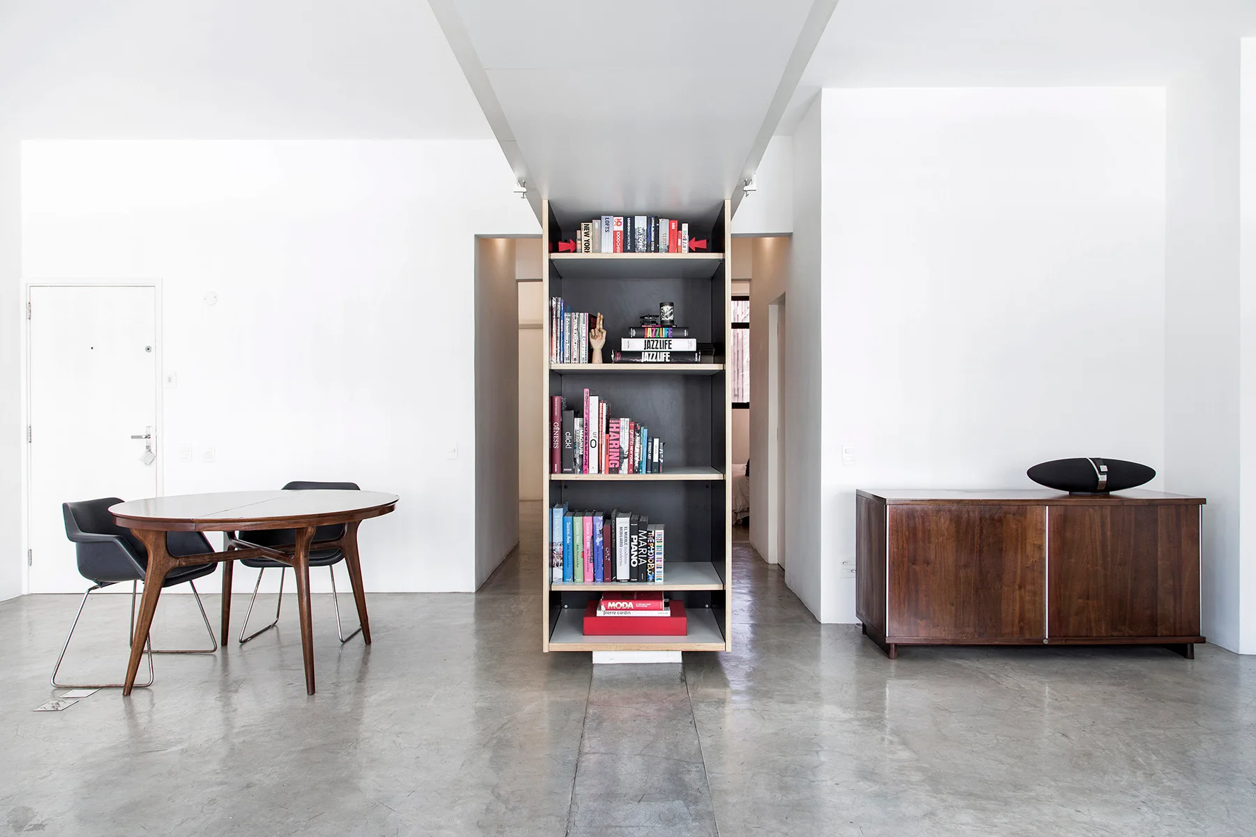A central wooden bookshelf divides two hallways in a minimalist room with a dining set, wooden credenza, and polished concrete floors.