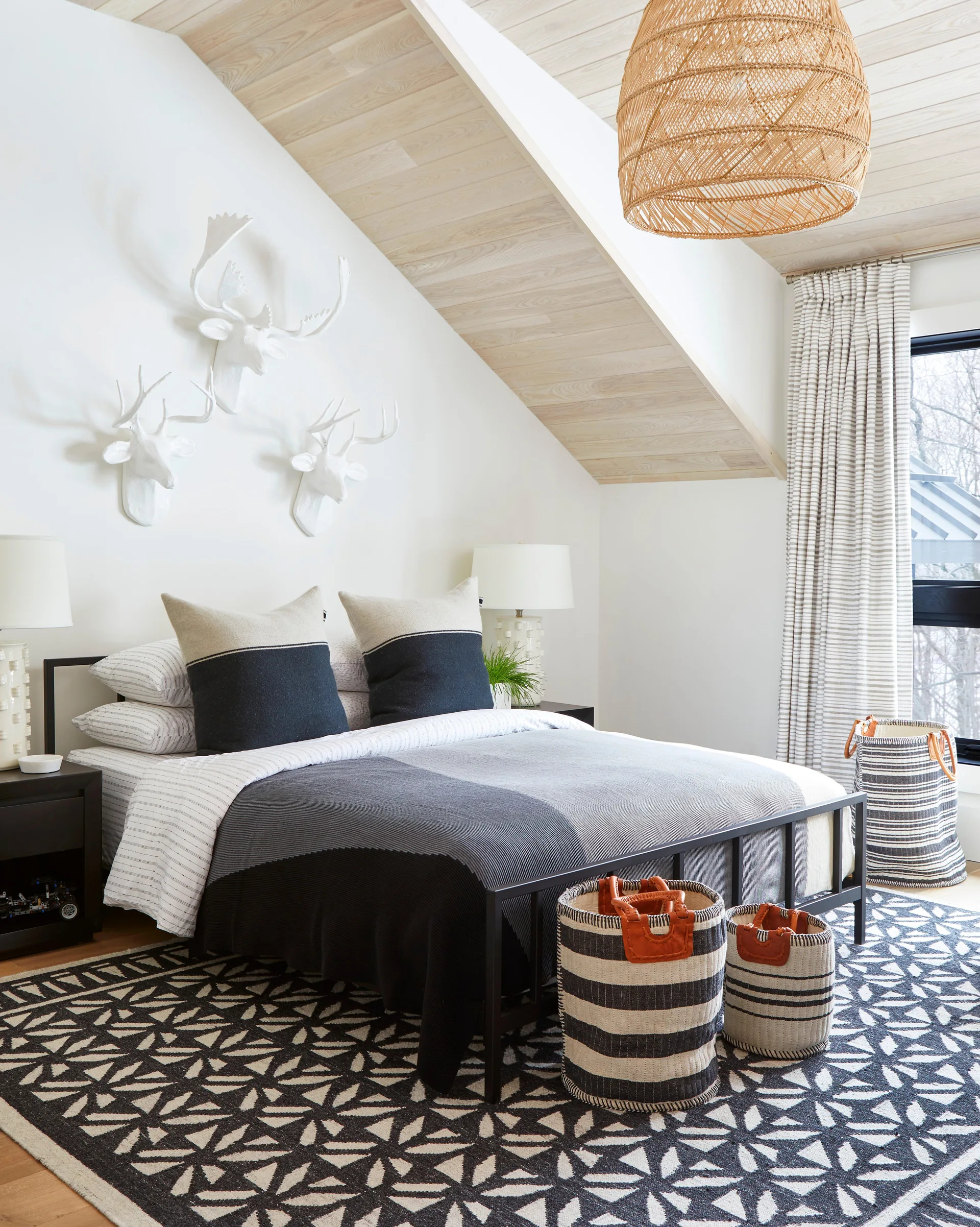A modern attic bedroom with a black metal bed, sloped wood-planked ceiling, a bold geometric rug, and white deer head sculptures on the wall.
