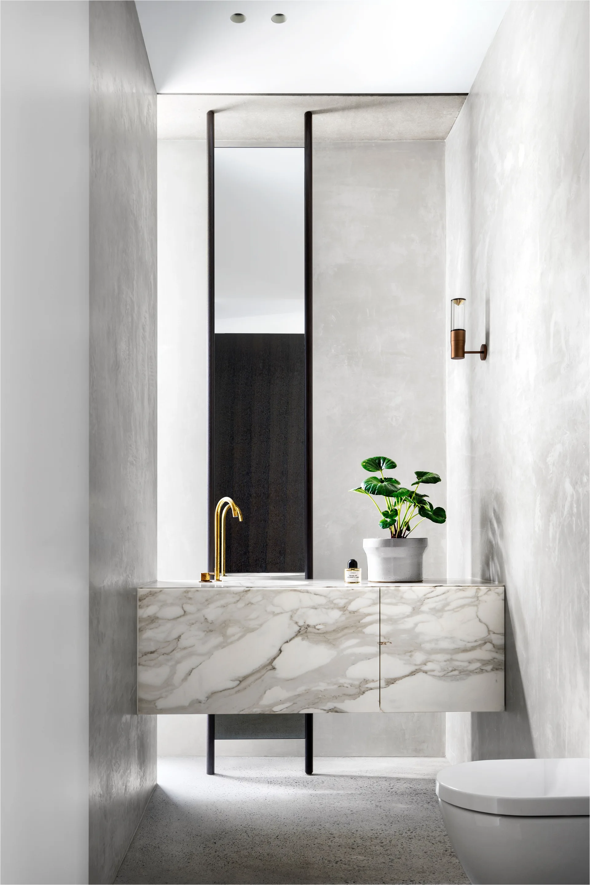 A modern powder room features a floating marble block vanity, curved brass faucet, textured plaster walls, and a tall vertical mirror.