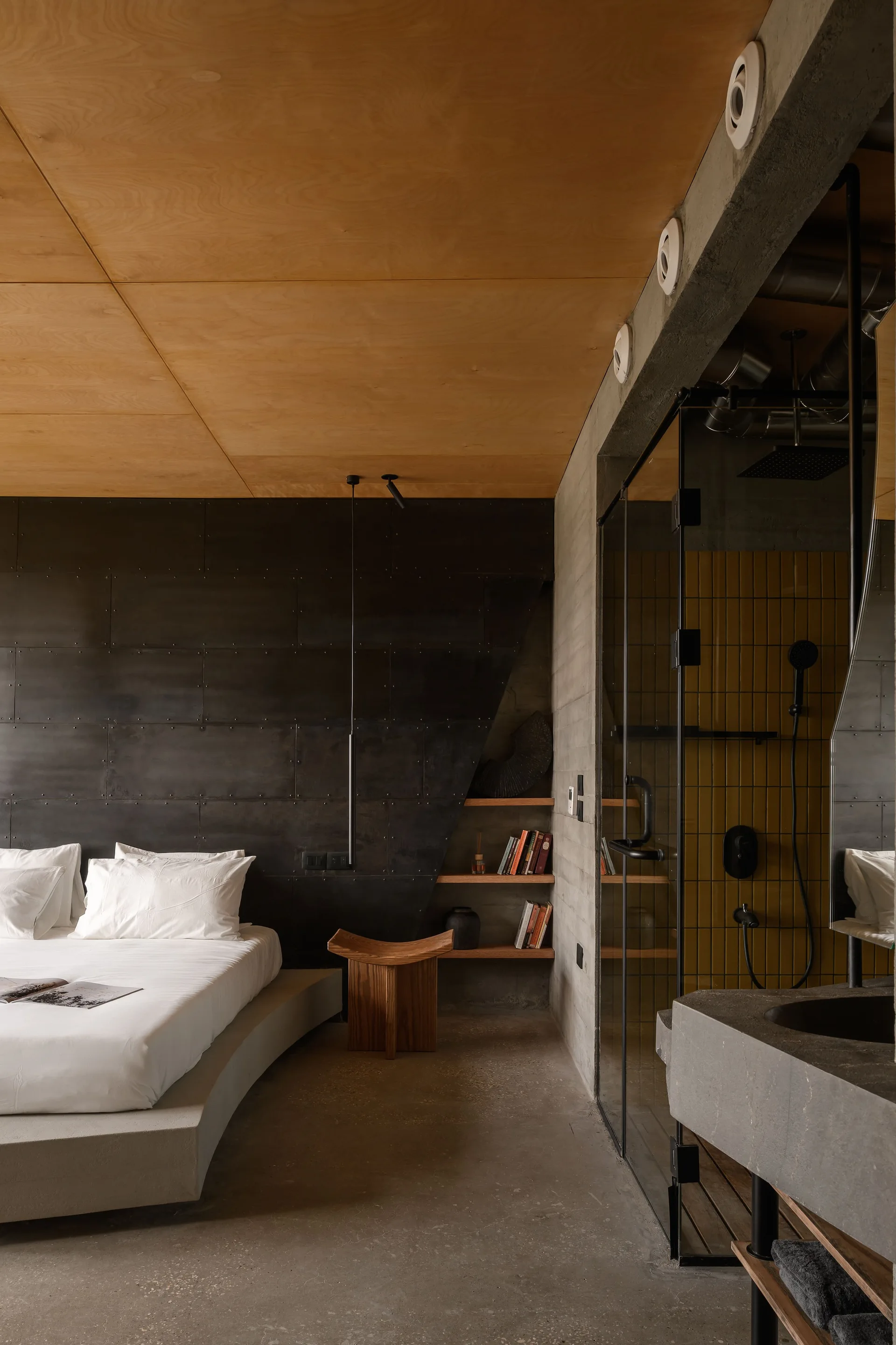 A modern industrial bedroom features a low platform bed against a black riveted metal wall beneath a warm plywood ceiling next to a glass shower.