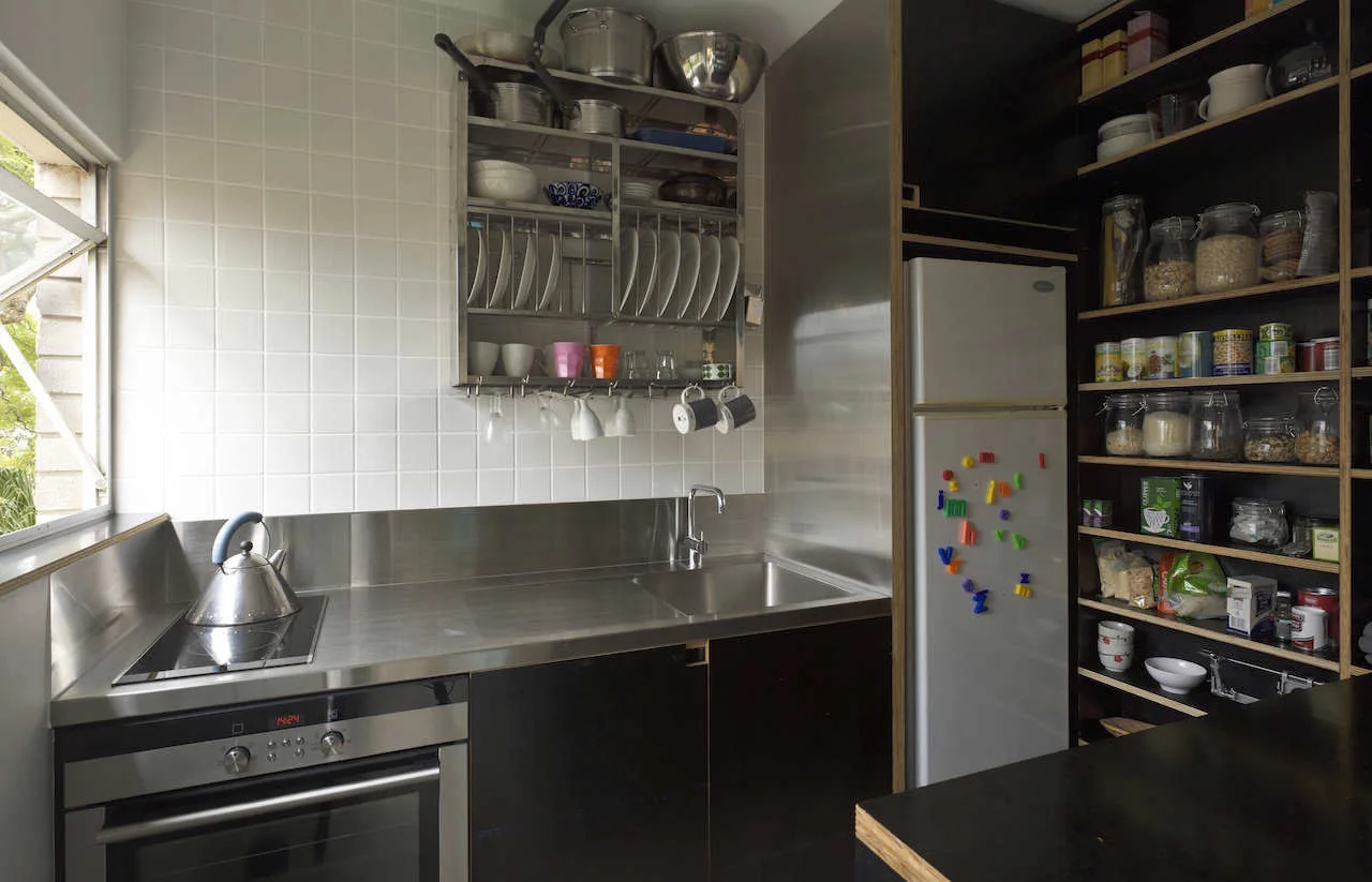 A compact kitchen featuring a stainless steel countertop with integrated sink, a grid tile backsplash, and dark open-shelving pantry.