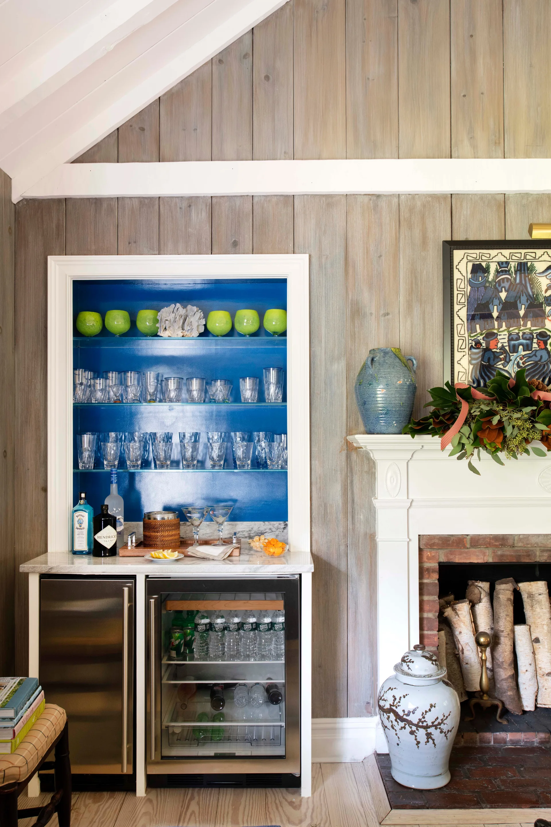A built-in home bar with a vibrant blue-painted shelf unit and an under-counter beverage fridge sits next to a brick fireplace with a white mantel.