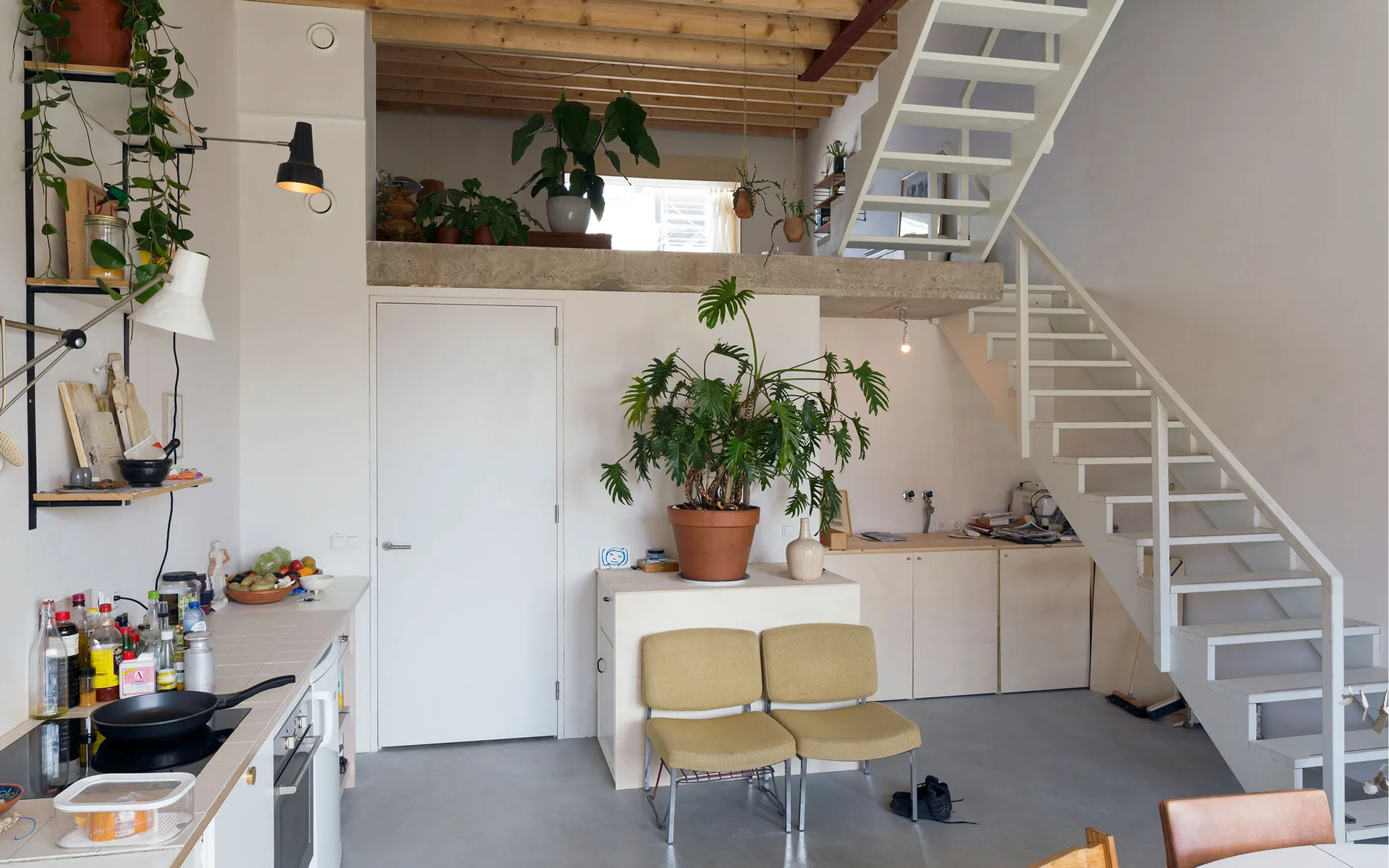 An open-plan loft featuring a white metal staircase next to light wood cabinets, topped by a mezzanine with exposed ceiling beams and potted plants.
