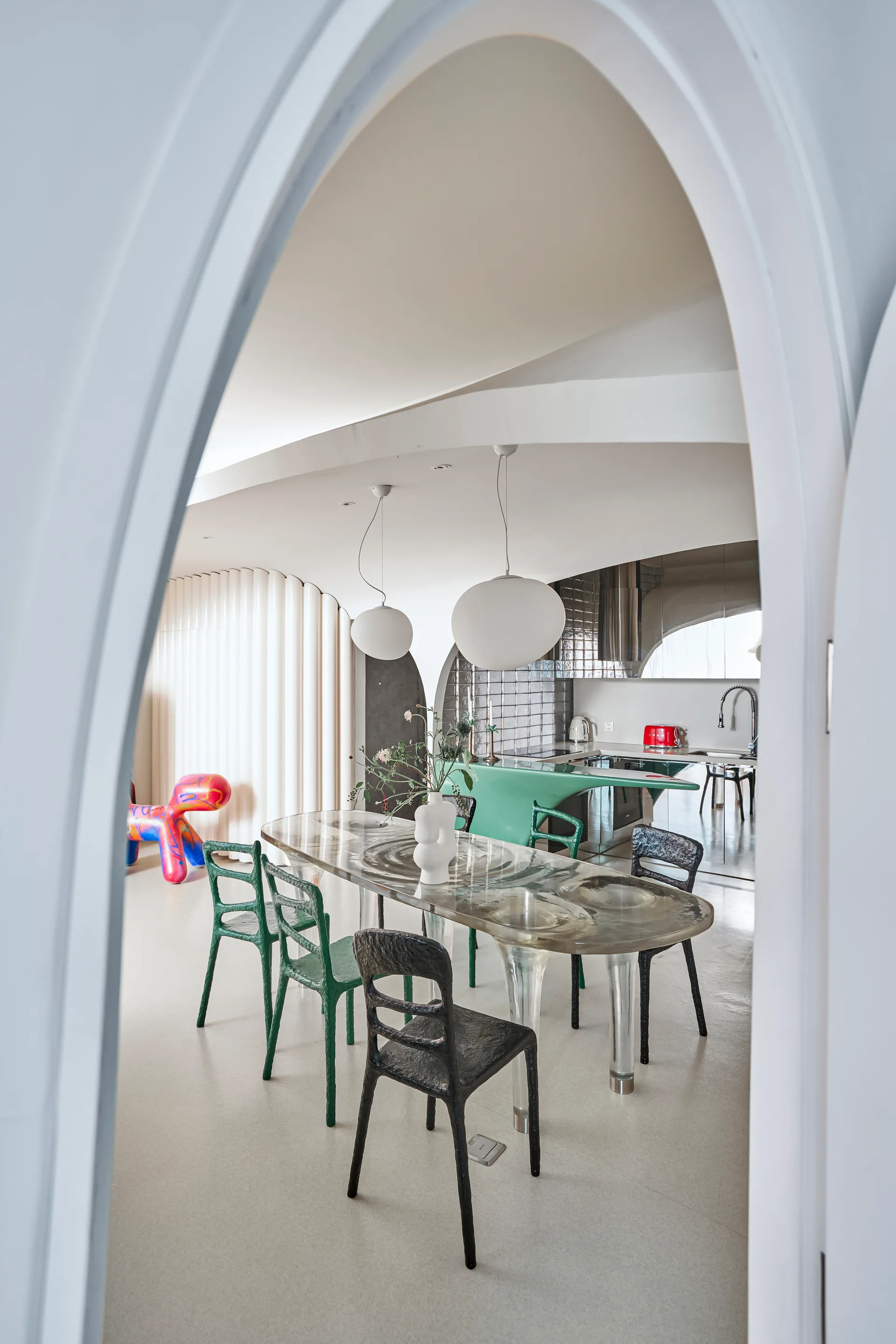 A contemporary dining room viewed through a pointed white archway, featuring a sculptural glass table, mismatched chairs, and globe pendant lights.