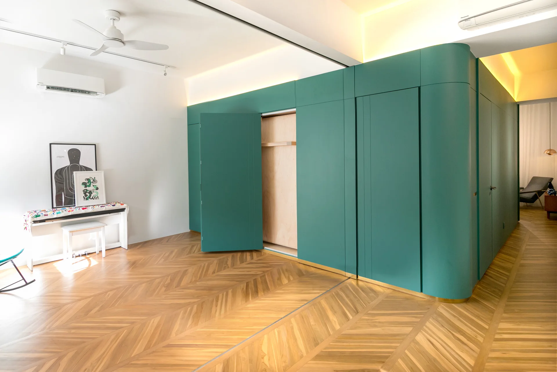 A spacious room featuring a curved matte teal storage wall with an open door, herringbone pattern wood flooring, and white walls with cove lighting.