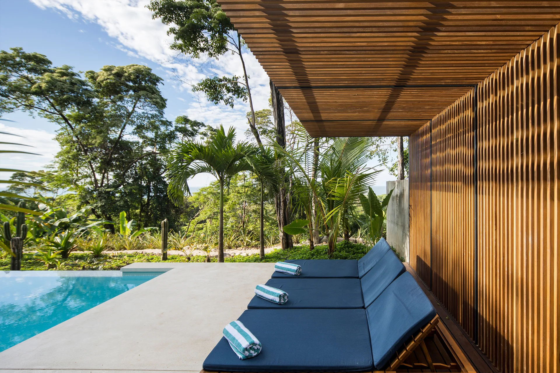 Blue cushioned lounge chairs sit on a concrete deck beside a swimming pool under a warm wood slatted canopy surrounded by tropical trees.