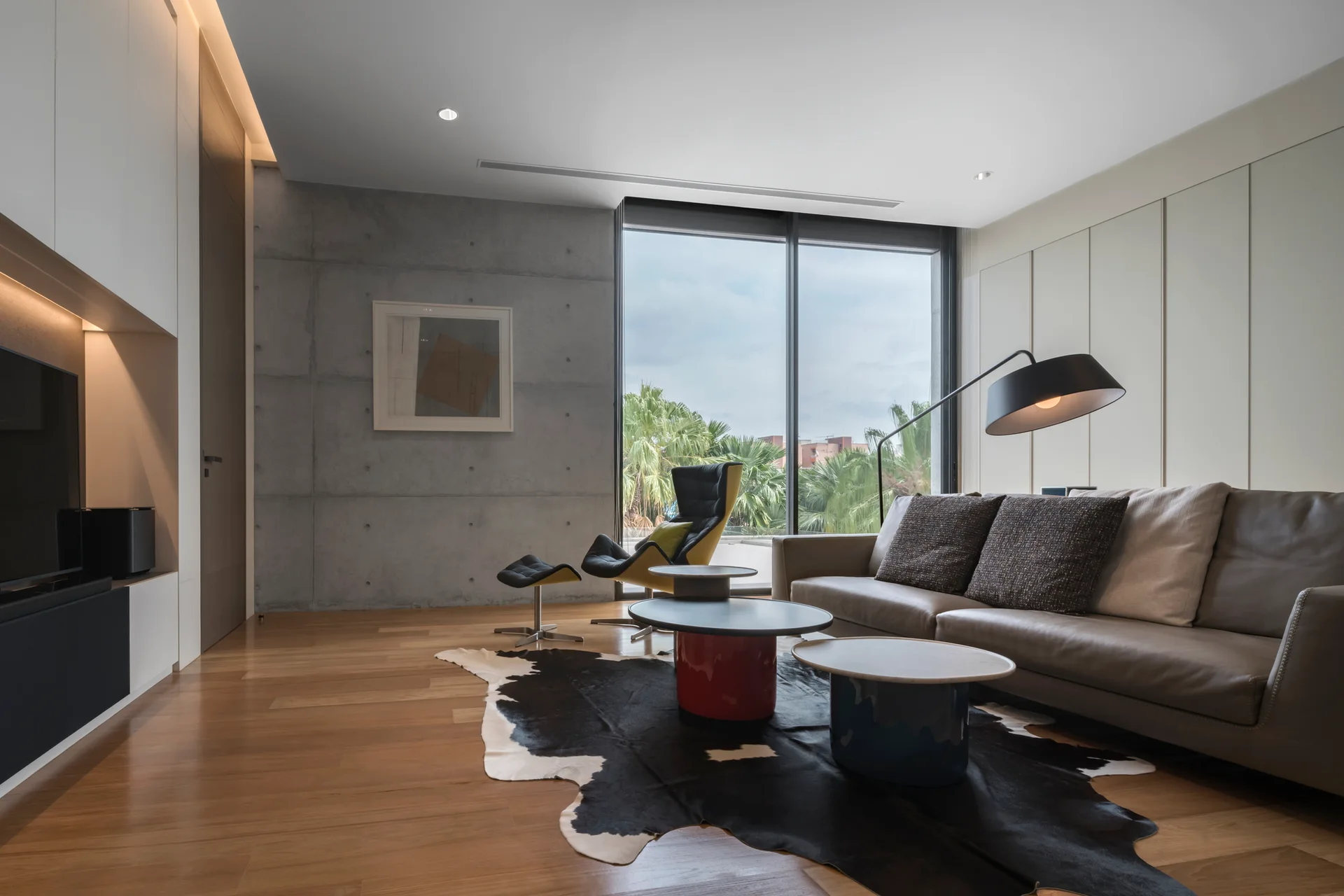 A modern living room features a leather sofa, a black-and-yellow lounge chair, and nested round coffee tables on a cowhide rug against a concrete wall.