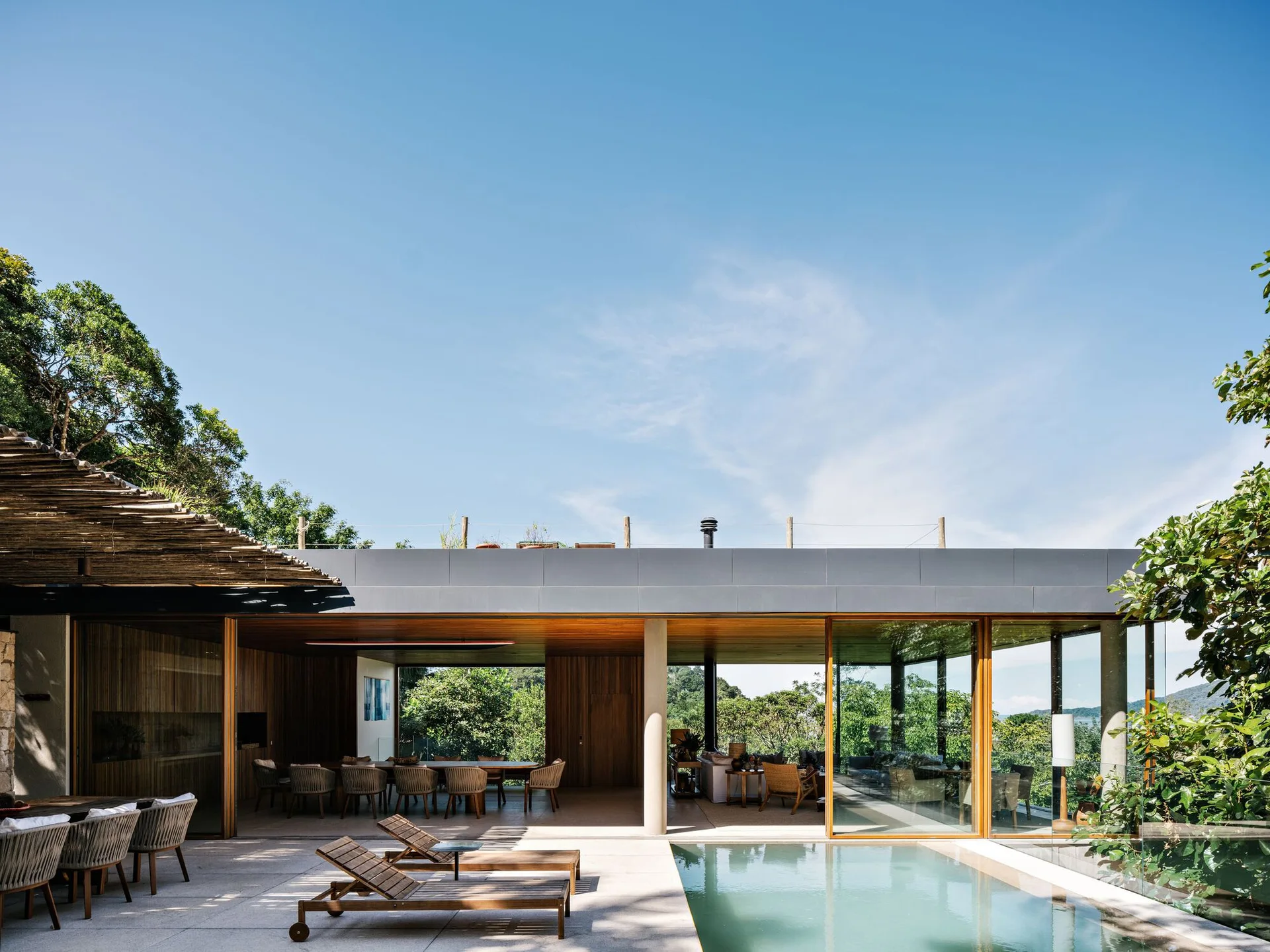 A modern stone patio features a swimming pool, wooden lounge chairs, and an open-air pavilion with glass doors and a warm wood ceiling.