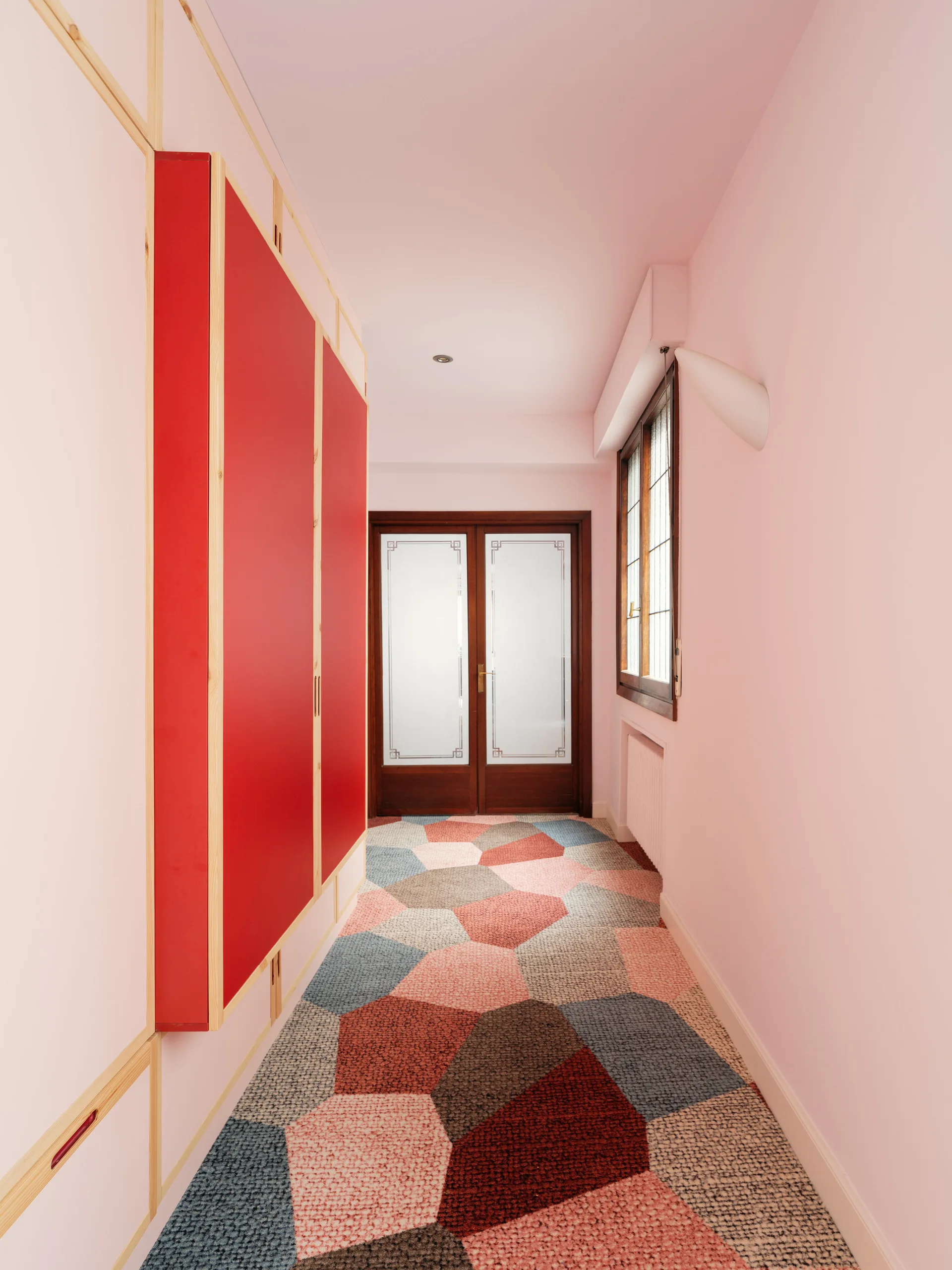 A narrow pink hallway featuring a large red built-in wardrobe with light wood trim and a multicolored geometric carpet.