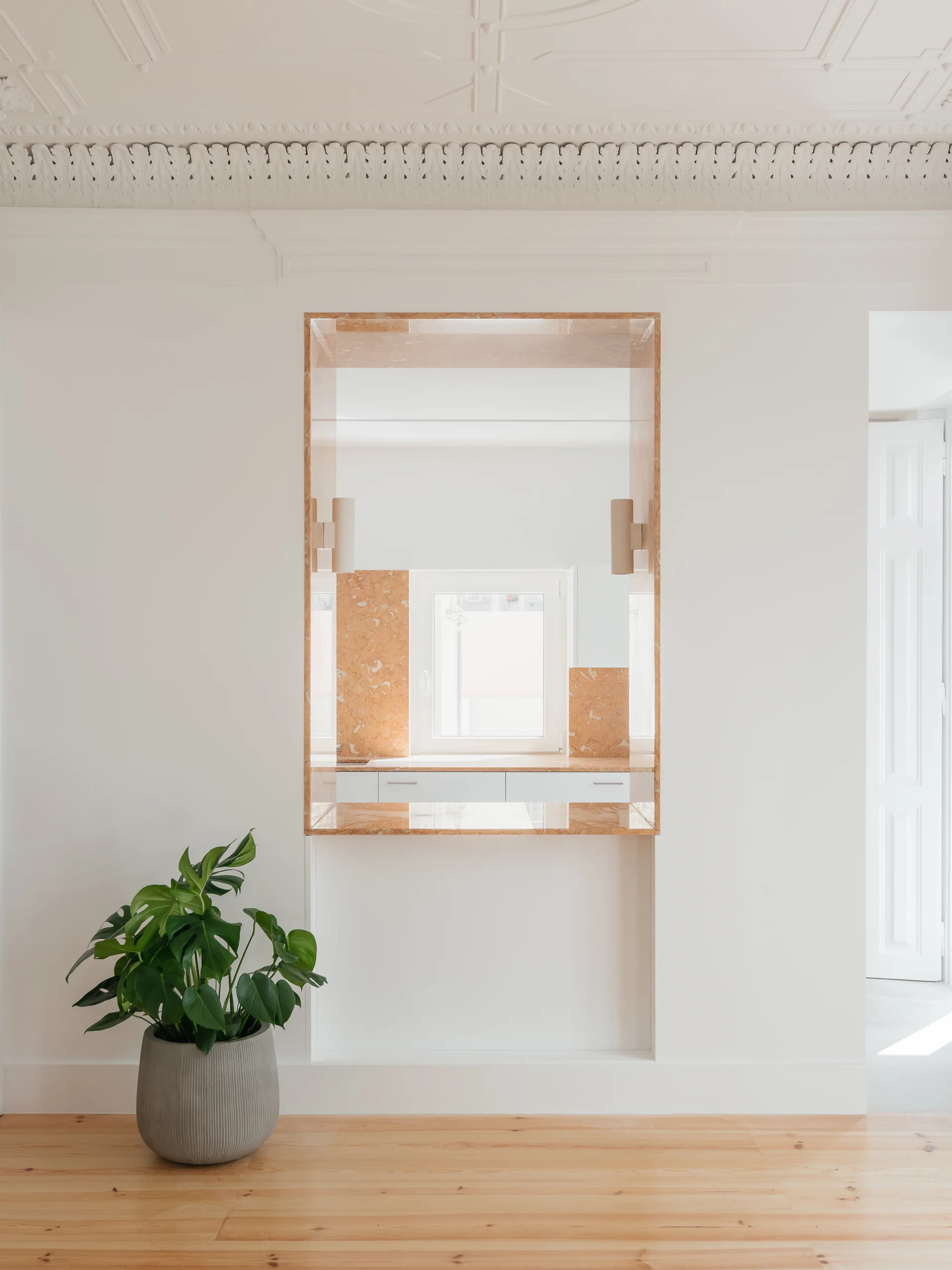 A white minimalist room features a central rectangular niche lined in peach marble framing a window, beside a potted monstera plant.
