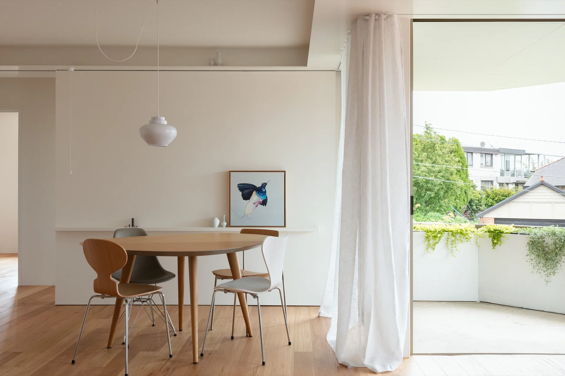 A light-filled dining room features a round wooden table, mixed modern chairs, a pendant light, and a sheer curtain opening to a balcony.