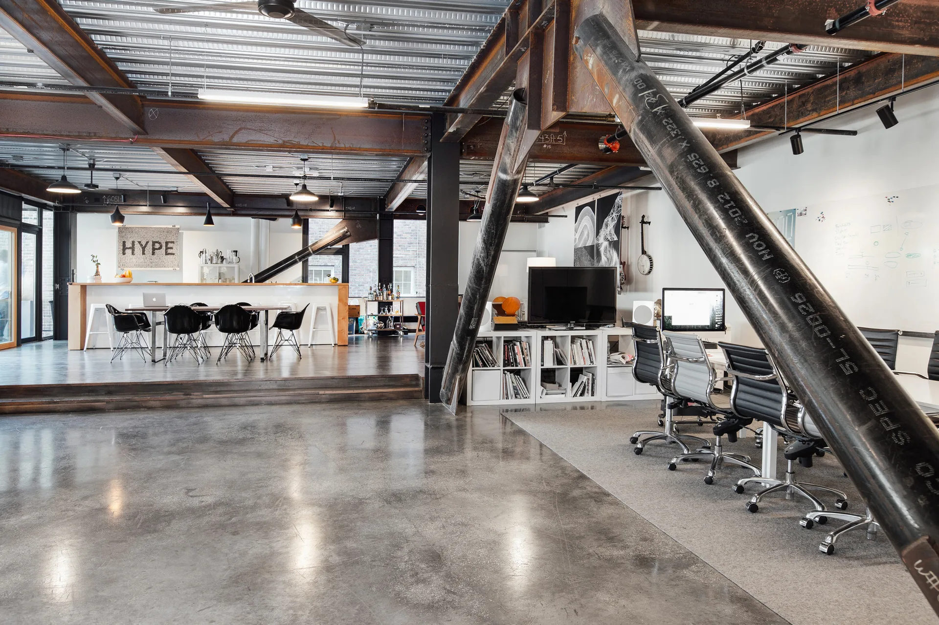 An industrial open-plan workspace featuring exposed structural steel beams, a polished concrete floor, office desks, and a raised kitchenette area.