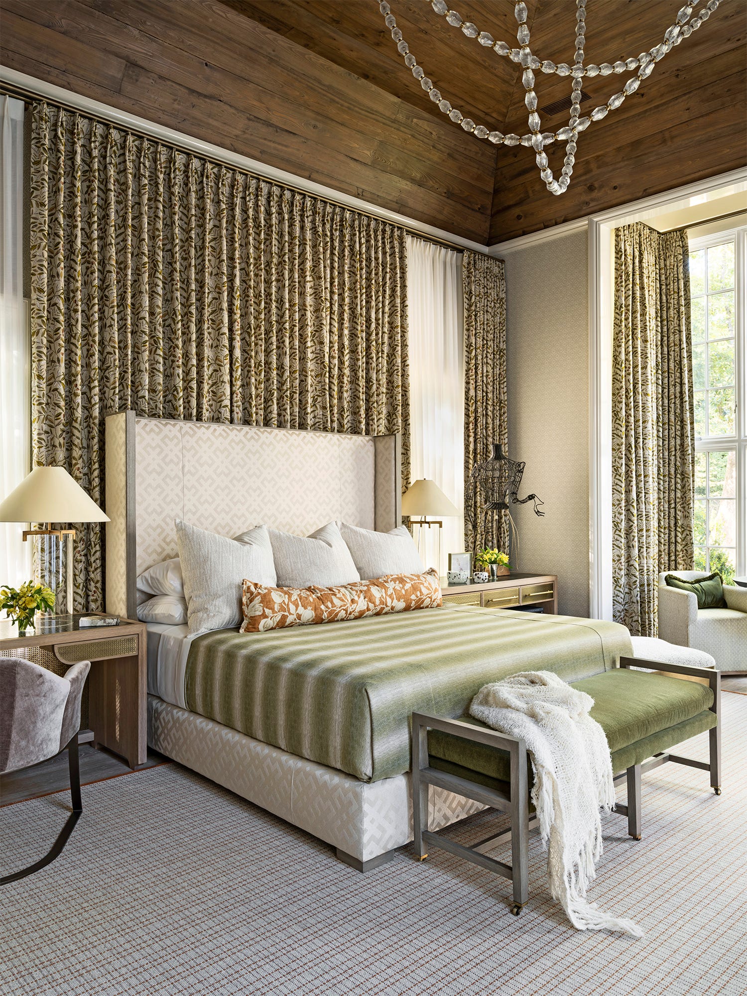 A sophisticated bedroom features a patterned upholstered headboard, floor-to-ceiling botanical curtains, and a dark wood plank ceiling.
