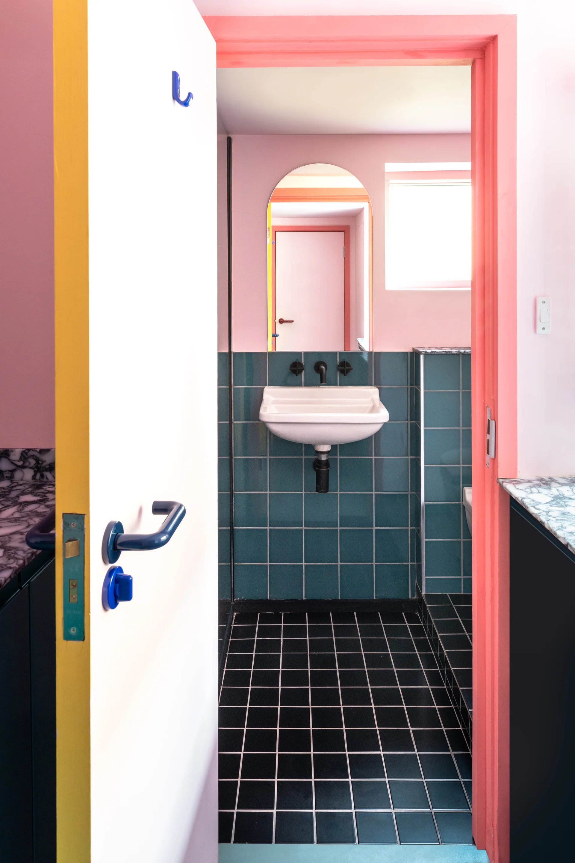 A view through a pink door frame reveals a bathroom with teal wall tiles, black floor tiles, a wall-mounted sink, and an arched mirror.