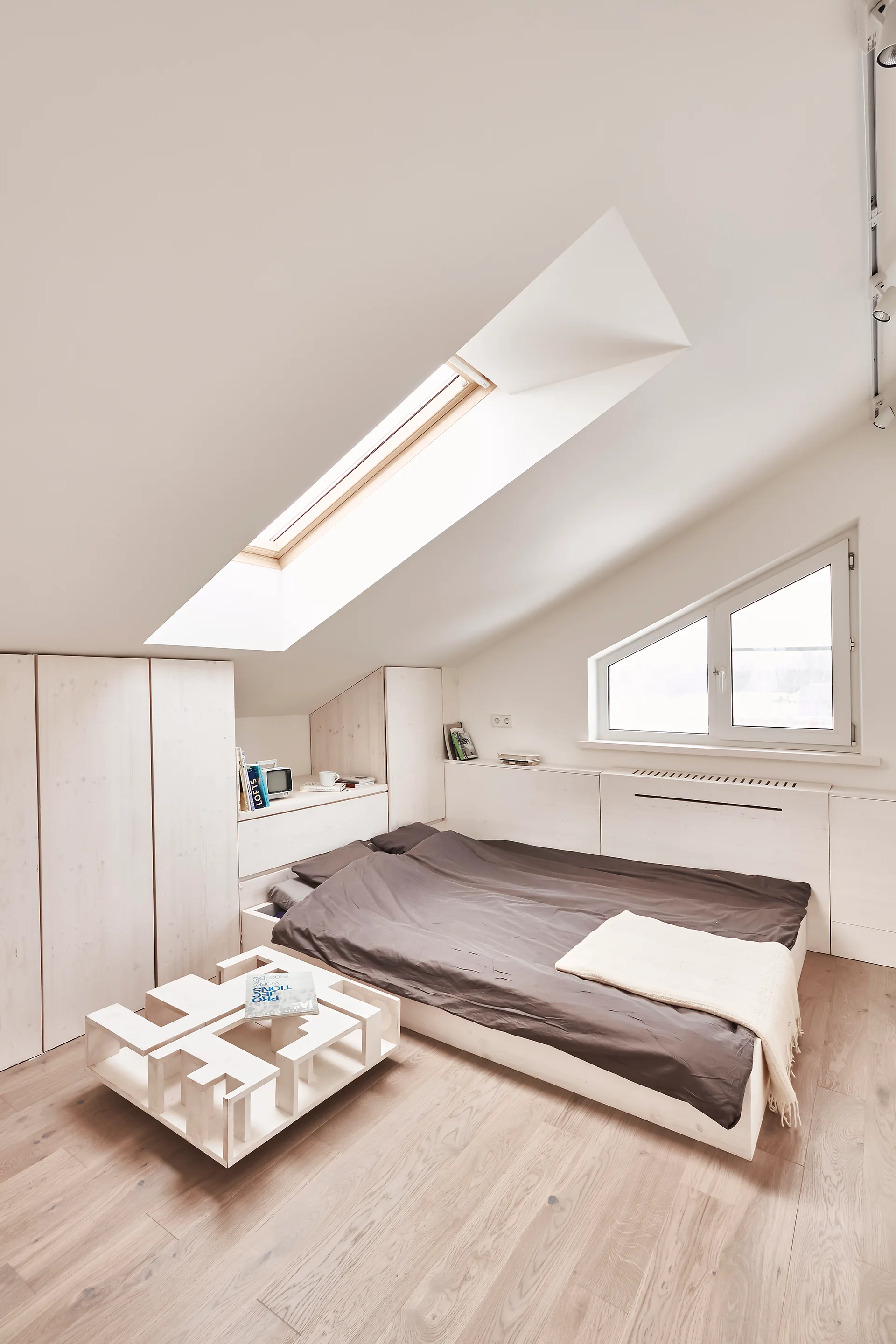 A minimalist attic bedroom features a low platform bed with dark bedding under a sloped ceiling with a skylight and custom built-in white cabinets.