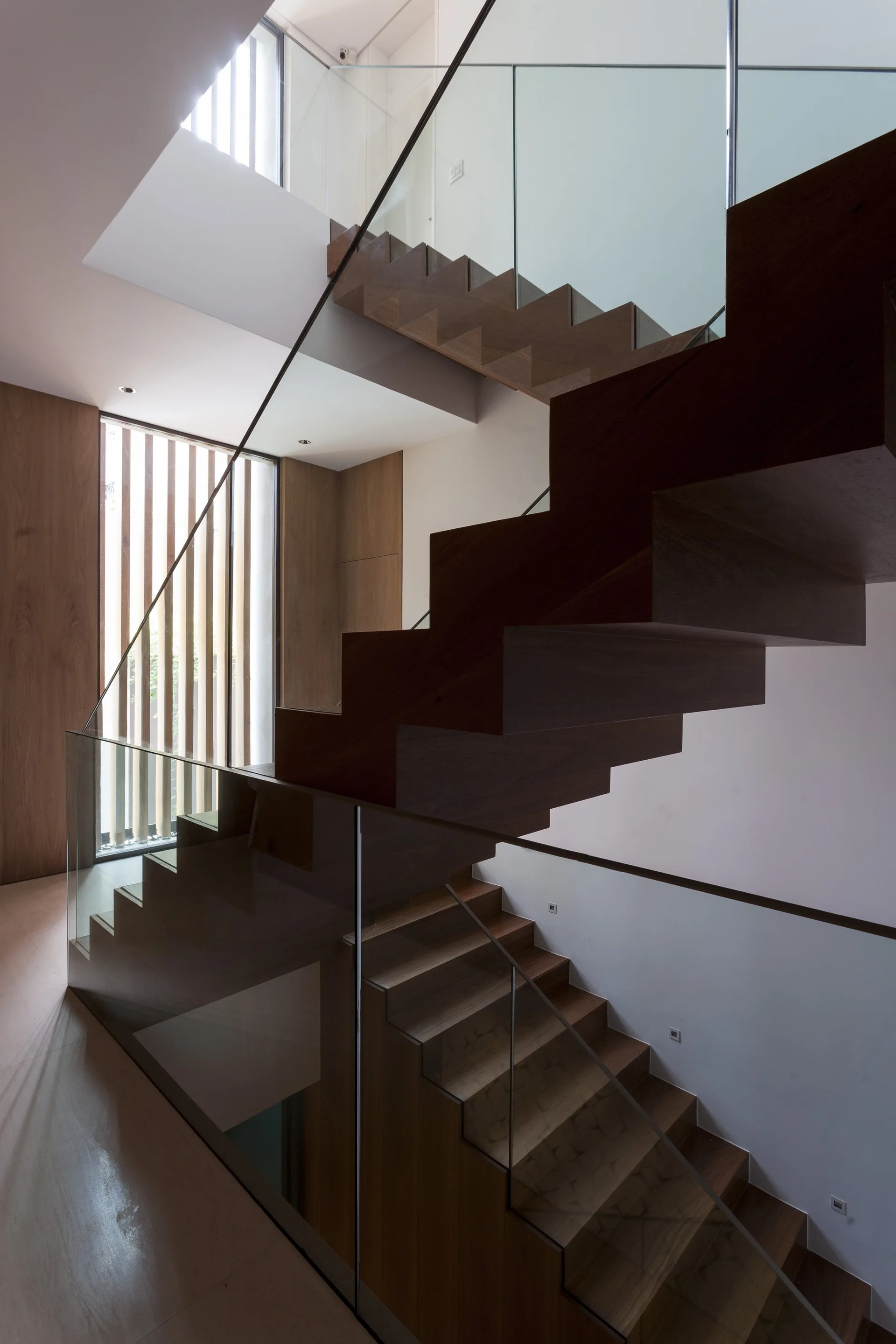 A modern multi-level staircase features floating dark wood treads, transparent glass railings, and a vertical wood slat window screen.