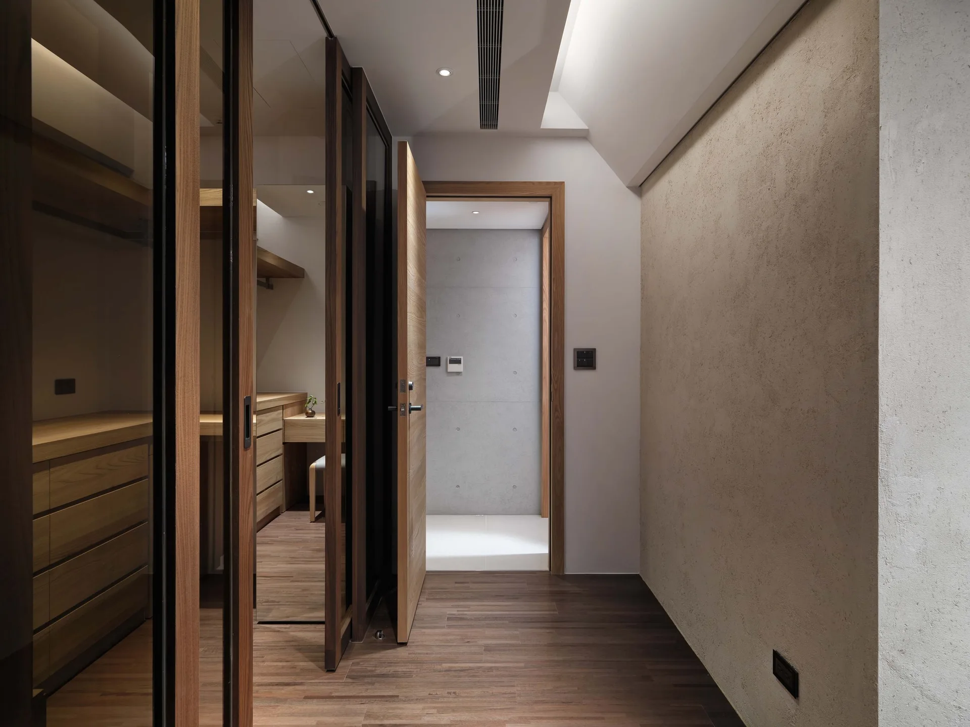 A modern hallway with wood floors and a textured beige wall leads past a glass-doored closet toward an open doorway.