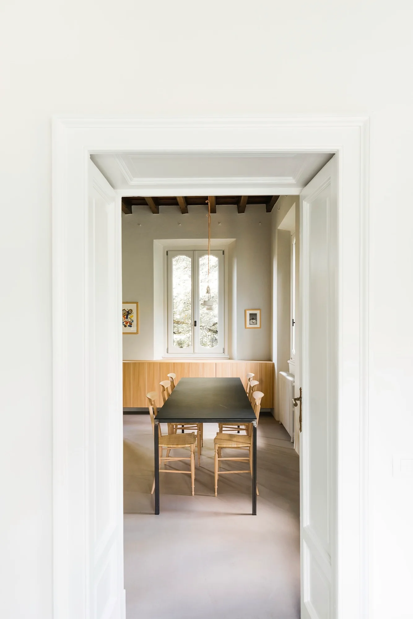 A view through a white doorway into a minimalist dining room featuring a black table, wooden chairs, and exposed timber beams.