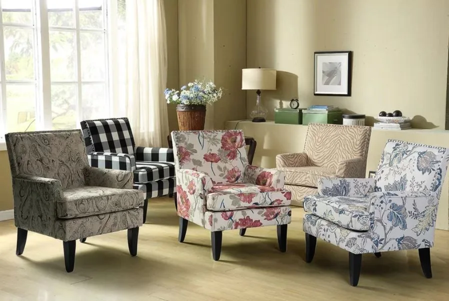 Five upholstered armchairs in various patterns including floral, checkered, and paisley are arranged on a light wood floor near a large window.