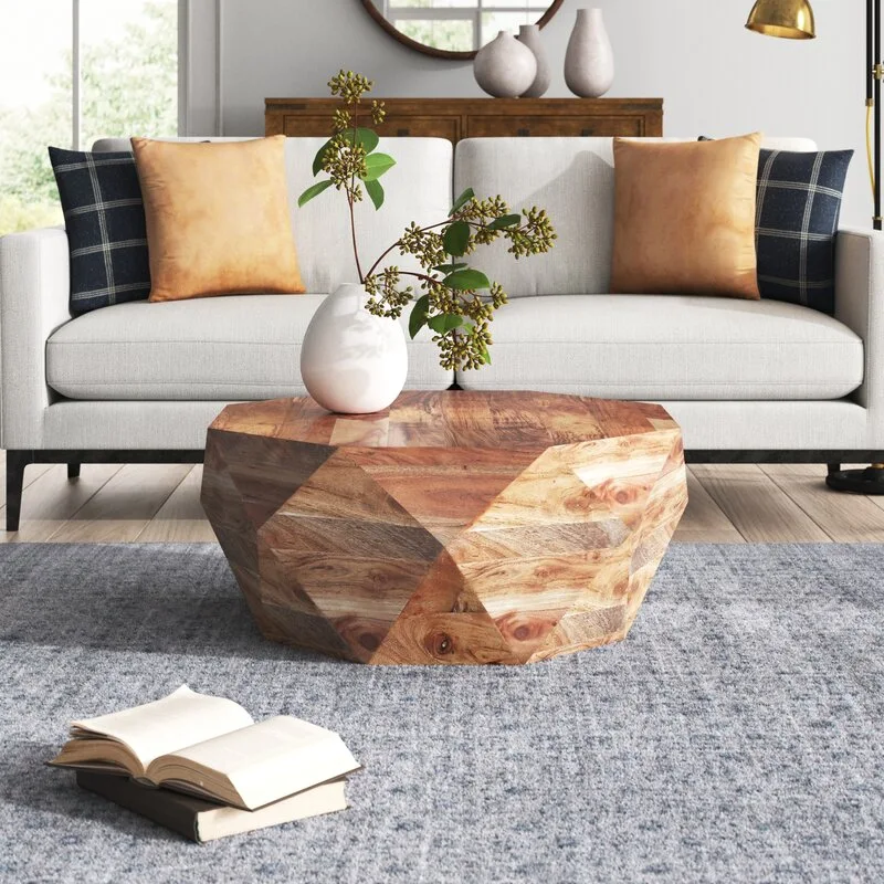 A faceted wooden coffee table with a white ceramic vase sits on a textured grey rug in front of a light grey sofa.