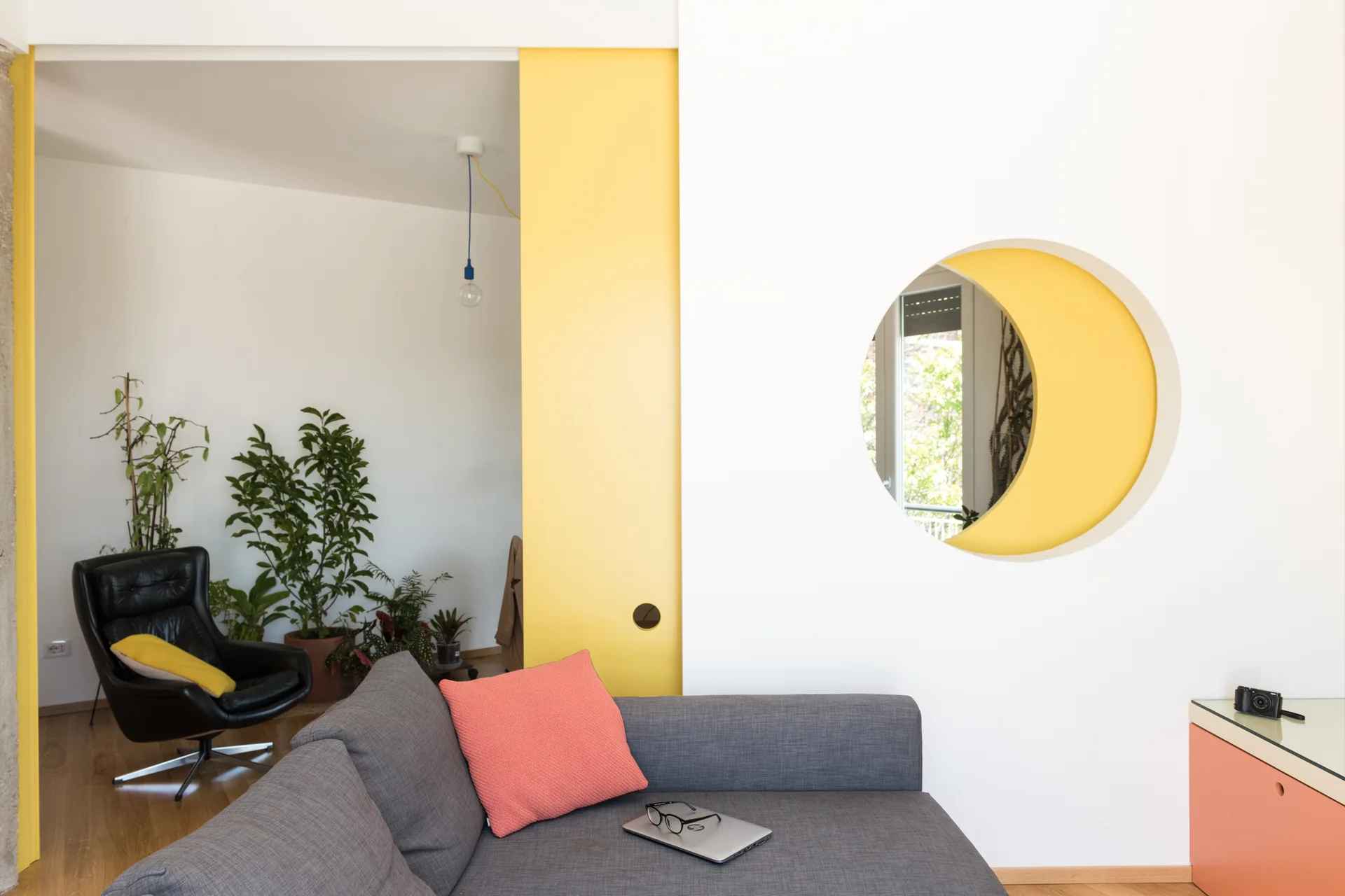 A modern living room featuring a grey sofa, a black leather armchair, and a white partition wall with a circular cutout revealing a yellow background.