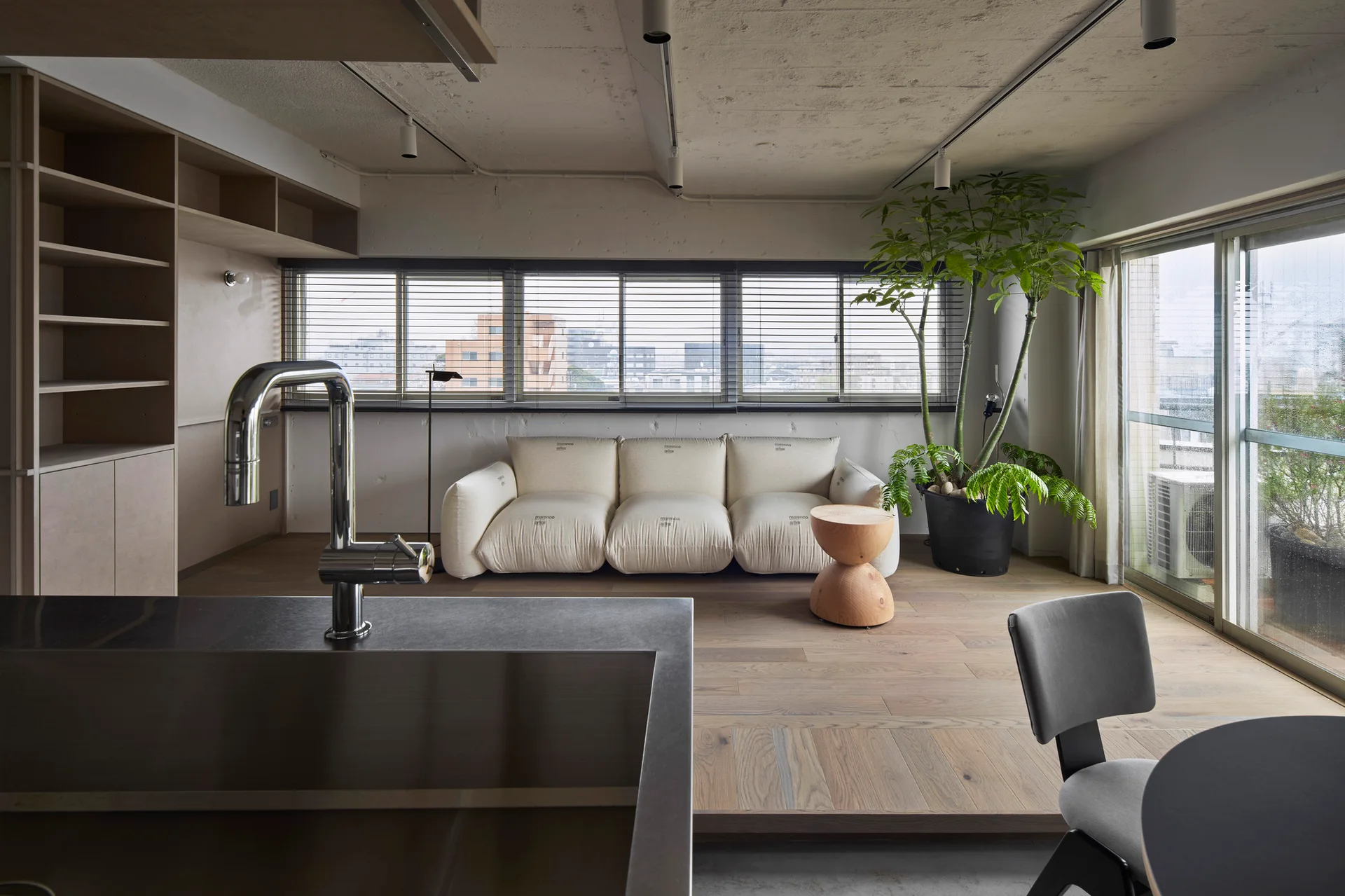 A living area with a cream sofa and wooden side table sits on a raised wood platform, next to large windows with blinds.