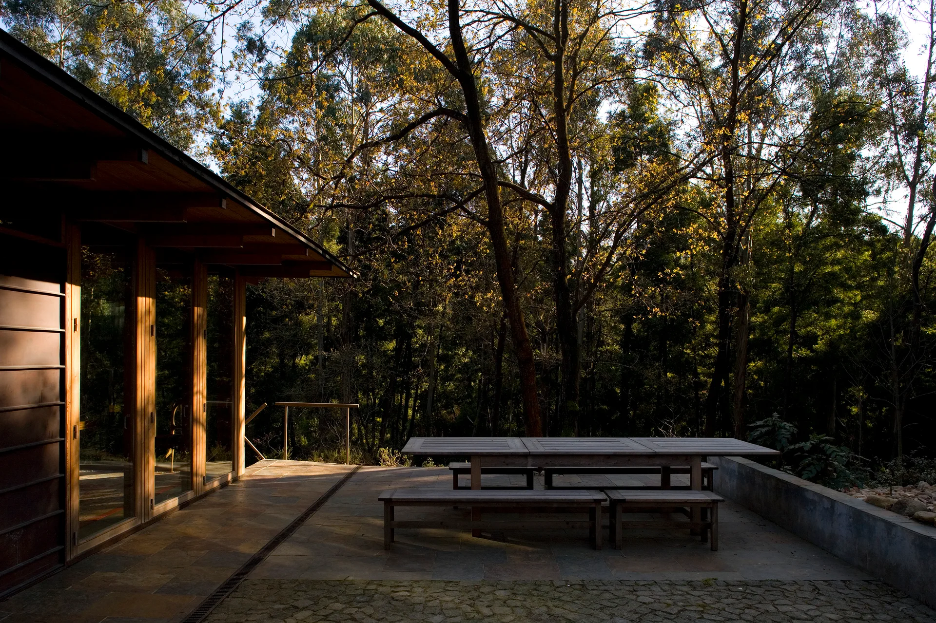 A stone patio features a long wooden dining table and benches next to a modern glass-fronted cabin surrounded by a dense forest.