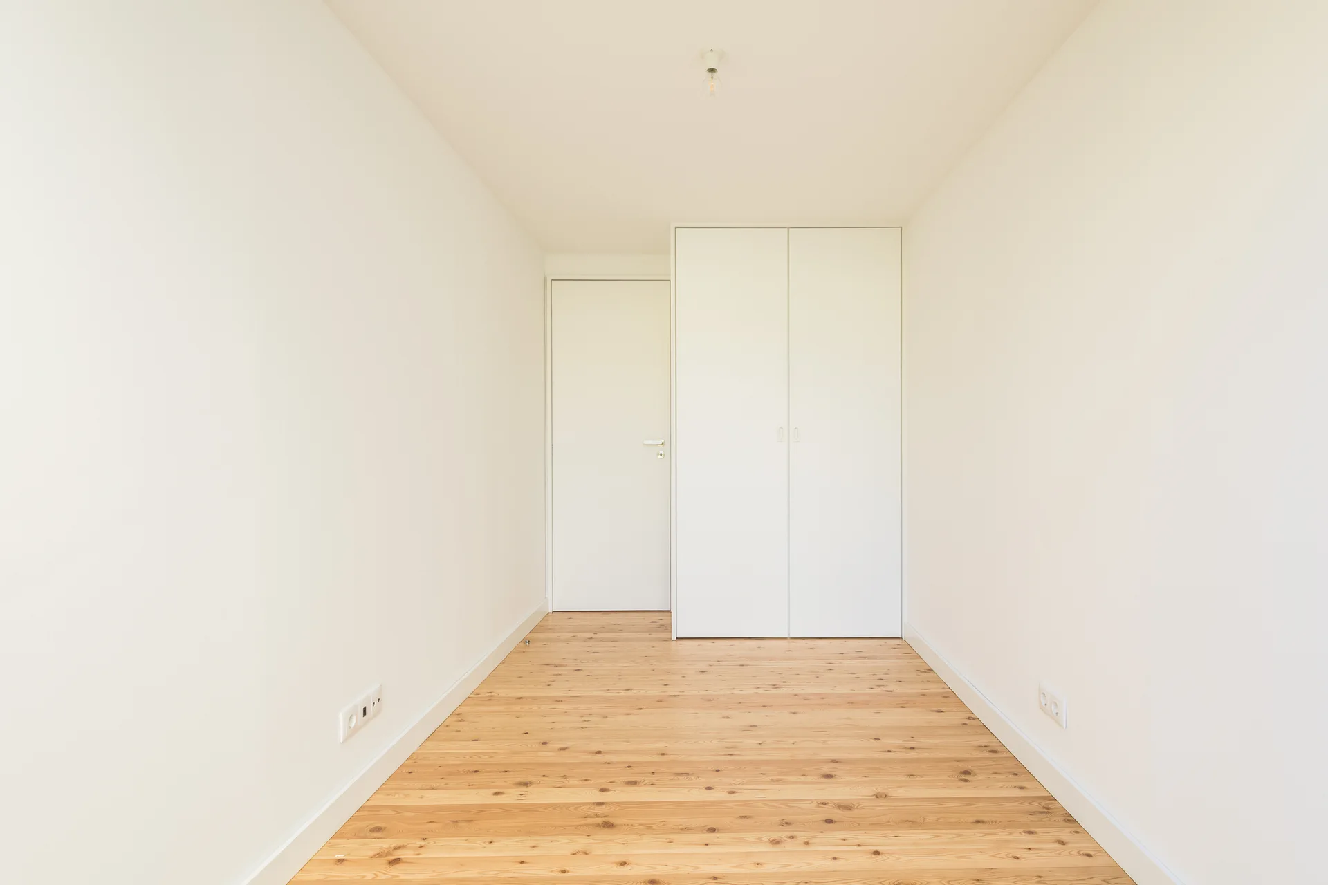 An empty minimalist room with light wood plank floors, clean white walls, a matching flush door, and a built-in wardrobe.