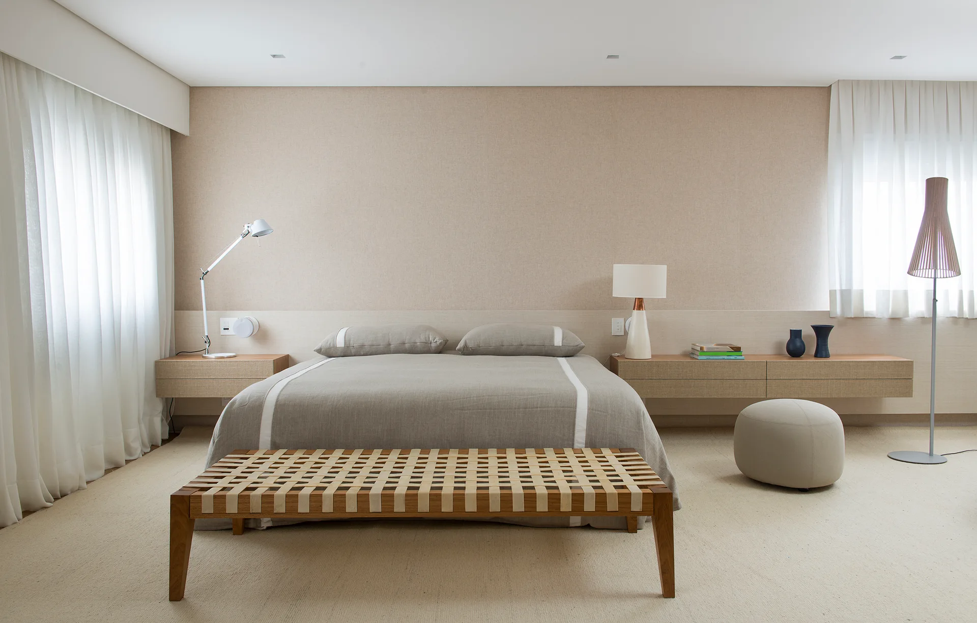 A minimalist bedroom features a central bed with grey linens, floating wooden nightstands, a woven strap bench, and sheer white curtains.