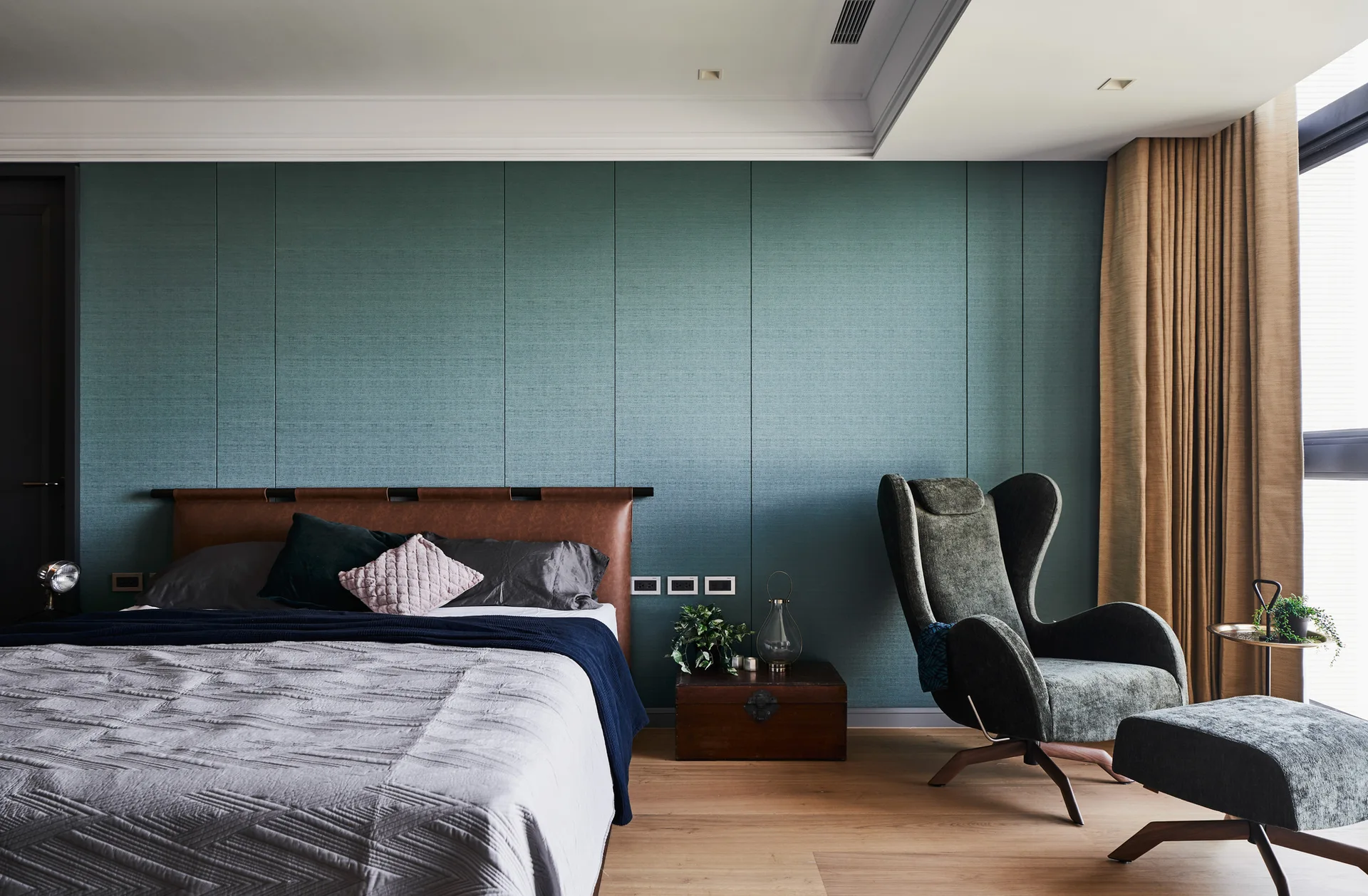 A modern bedroom features a textured teal paneled wall behind a bed with a brown leather headboard and a dark grey armchair with an ottoman.