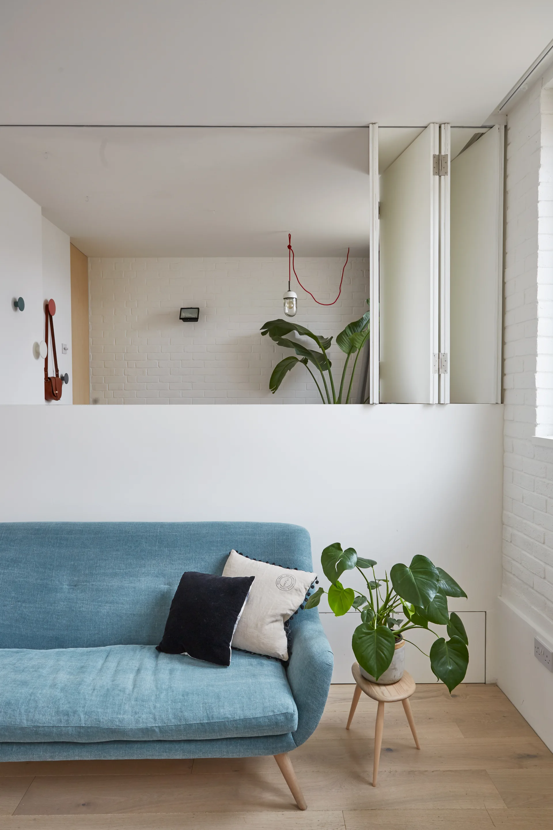 A blue fabric sofa sits against a white partition wall next to a potted plant on a wooden stool, with a folding door opening to an upper room.