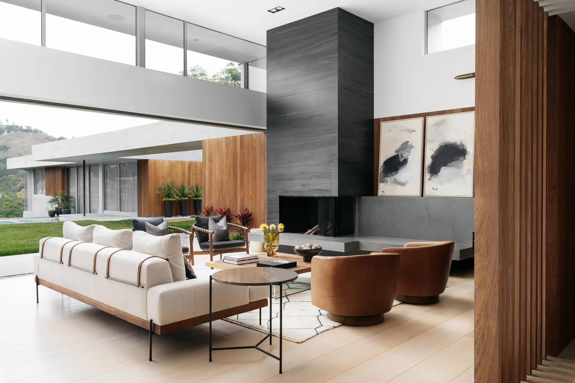 A modern open-plan living room featuring a cream sofa, tan leather armchairs, a large dark fireplace column, and a wide open wall leading outside.