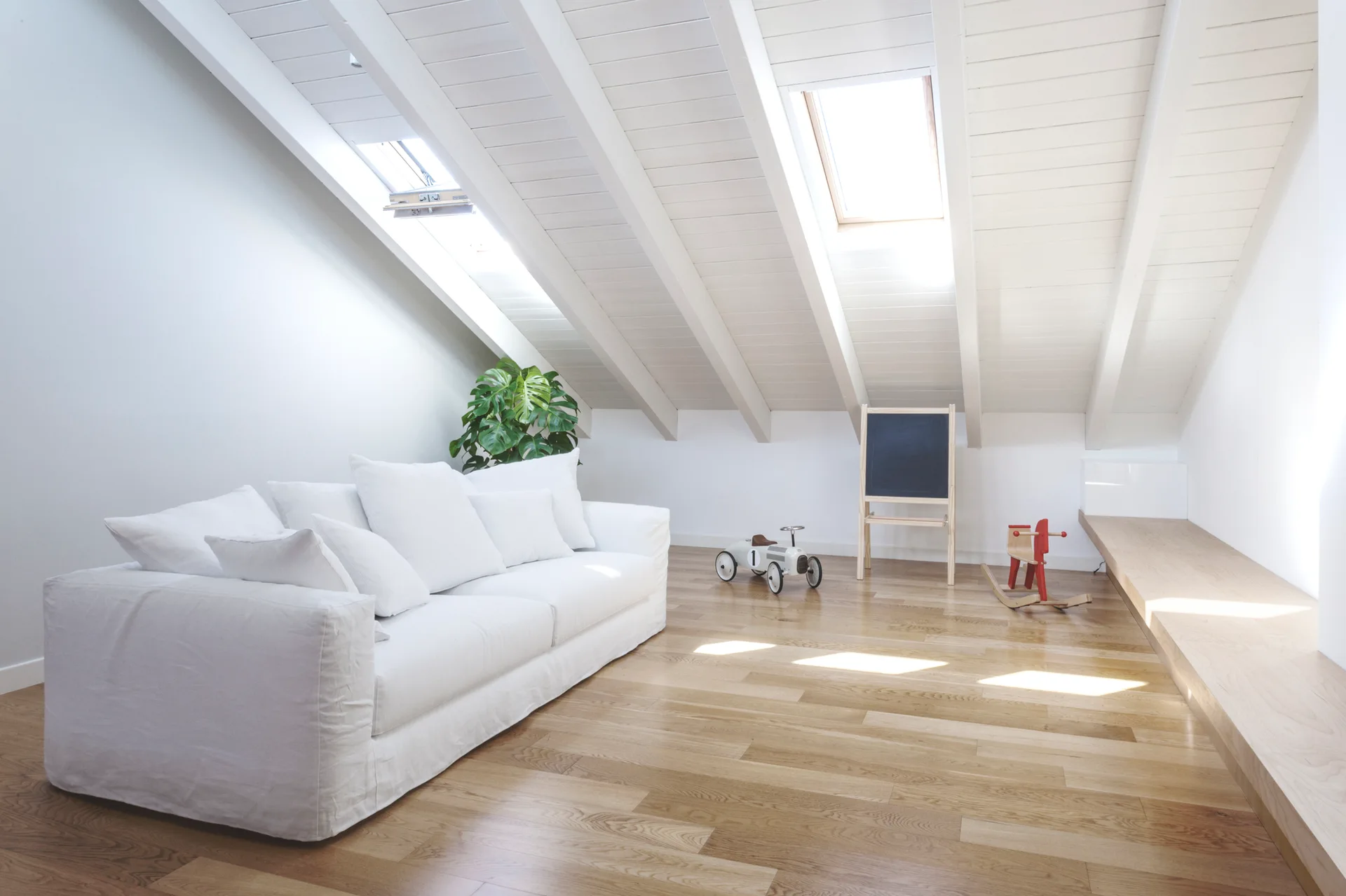 A bright attic playroom featuring a white sofa, sloped ceilings with skylights, wooden floors, a low wood bench, and children's toys.