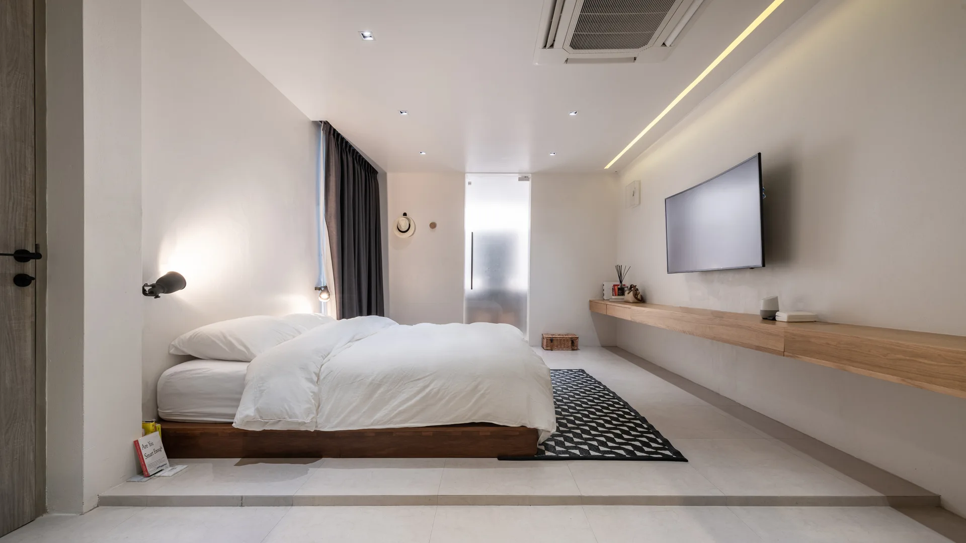 A minimalist bedroom featuring a low wooden platform bed on a raised floor, a long floating oak console, and a wall-mounted television.
