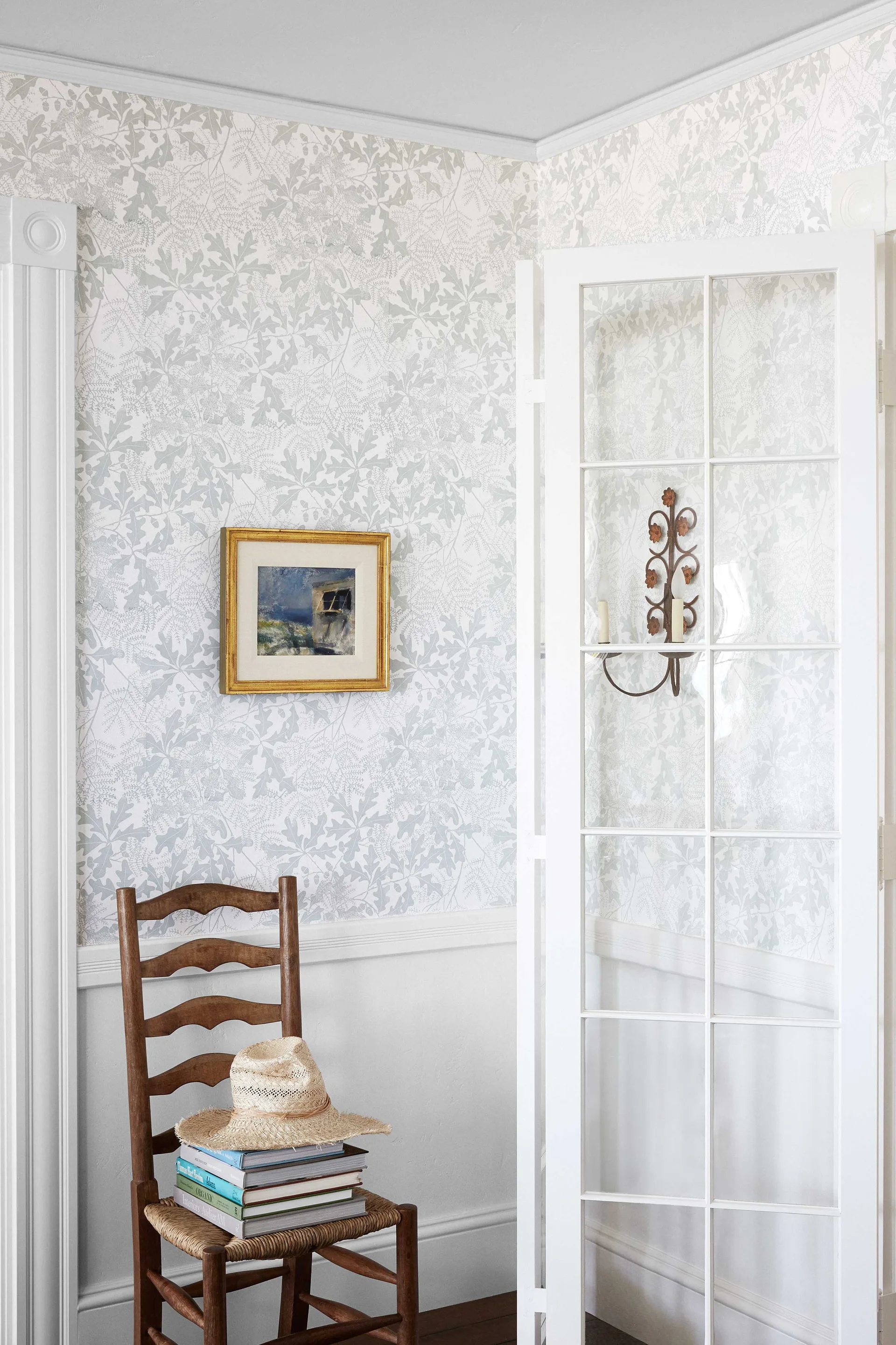 A wooden ladder-back chair with a straw hat sits against white wainscoting and botanical wallpaper next to a glass-paneled French door.