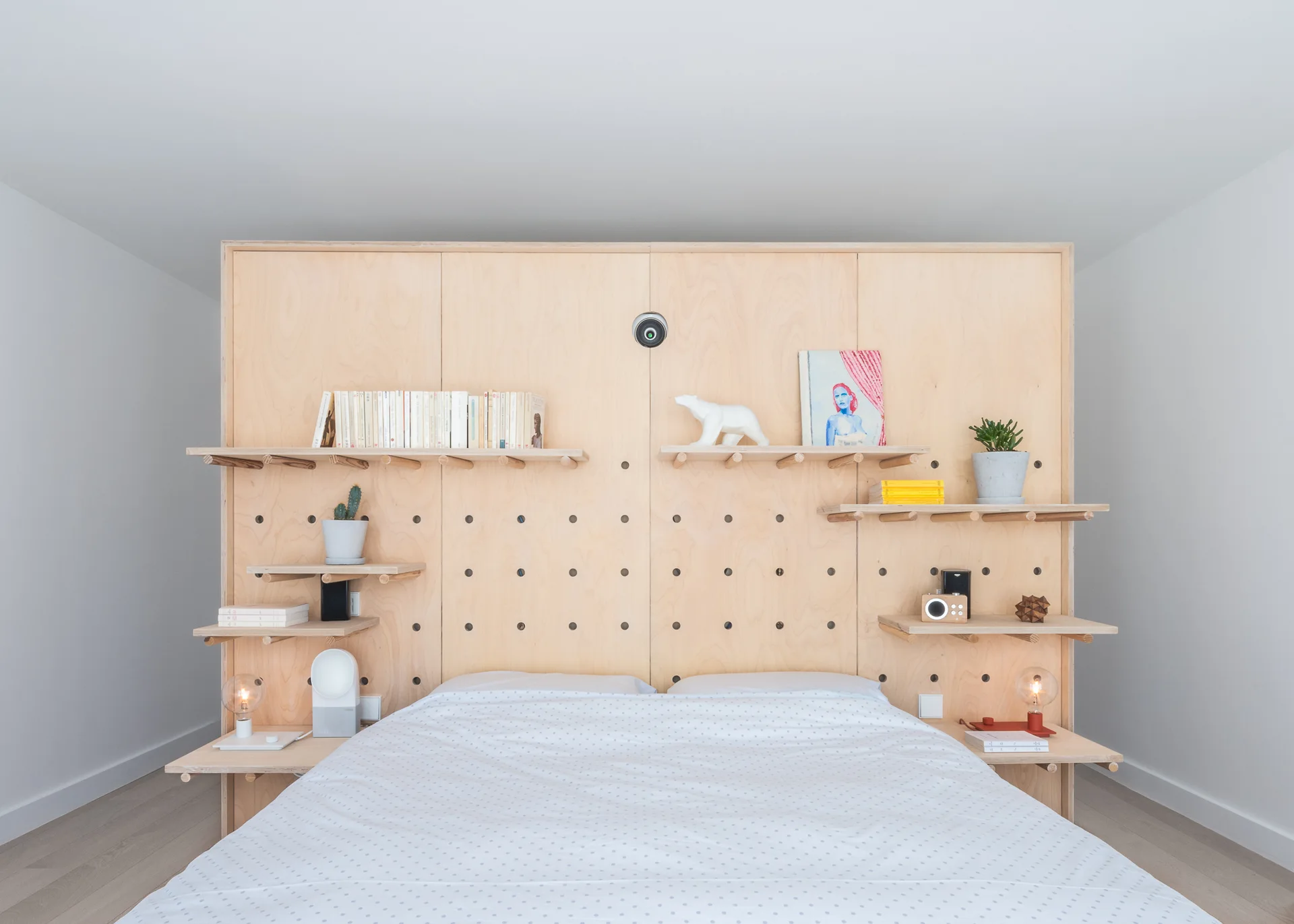 A bed with white bedding stands against a large plywood pegboard headboard with floating shelves holding books, small plants, and decor.