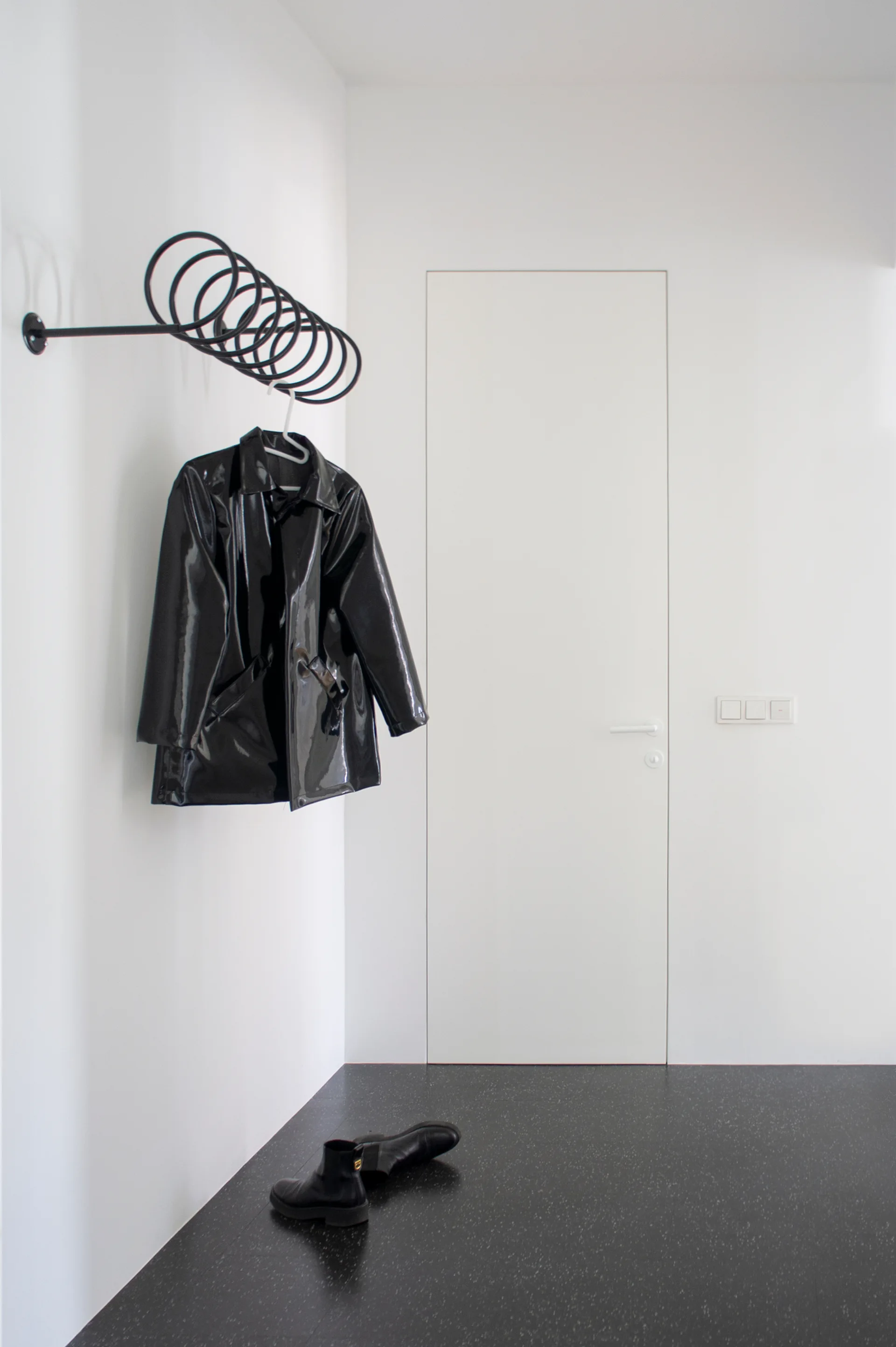 A black spiral metal coat rack holds a glossy black jacket in a minimalist white hallway with a flush white door and dark flooring.