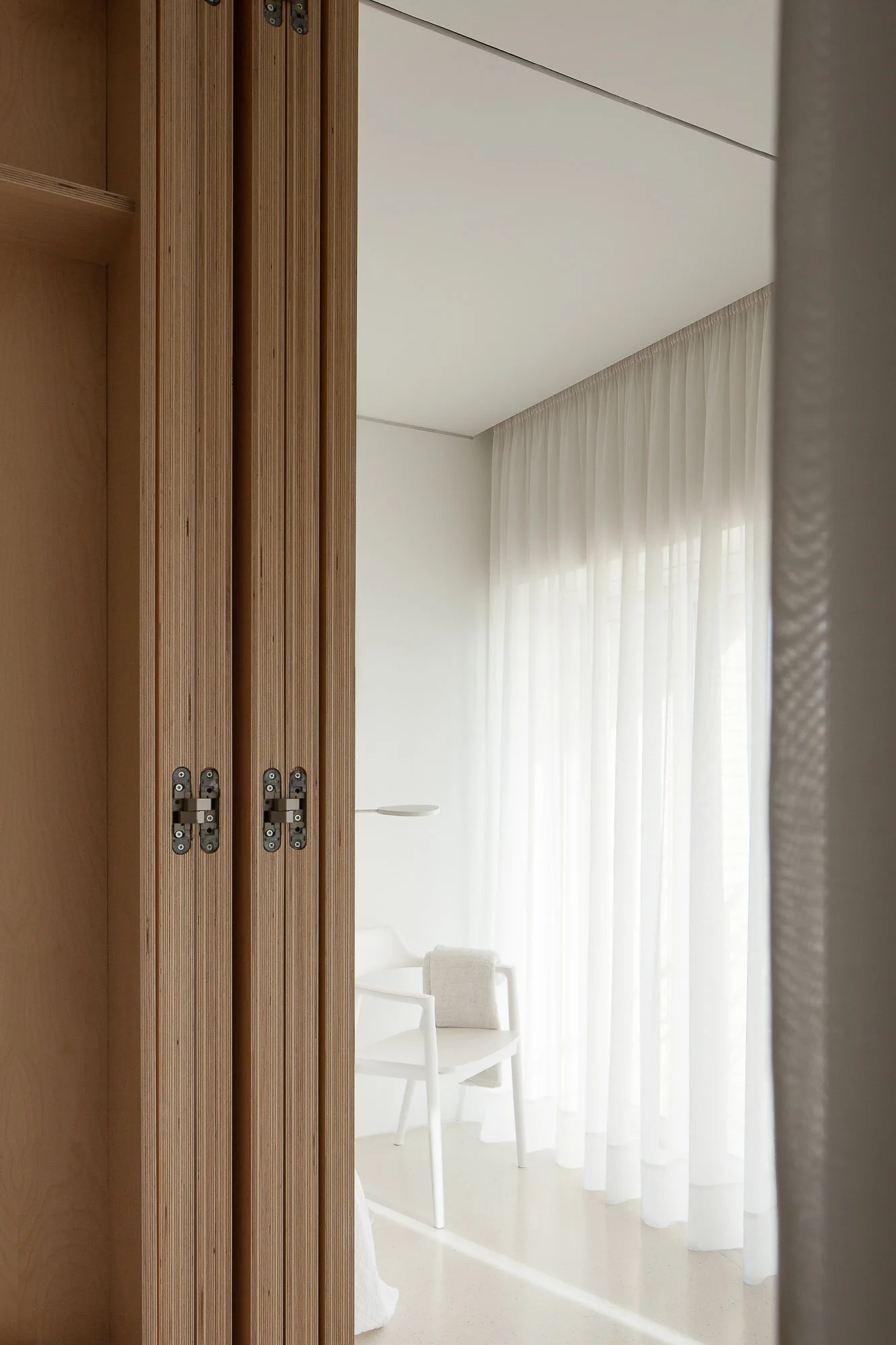 A view through a light wood plywood folding door with exposed hinges into a bright white bedroom with sheer curtains and a chair.