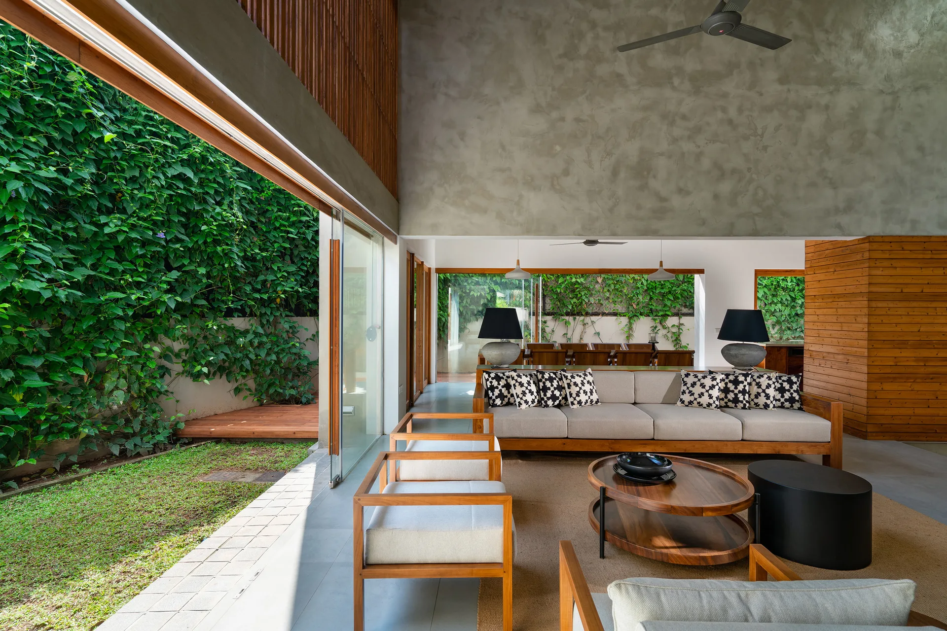 A modern living room features wooden furniture, a concrete feature wall, and large sliding glass doors opening to a lush green garden.
