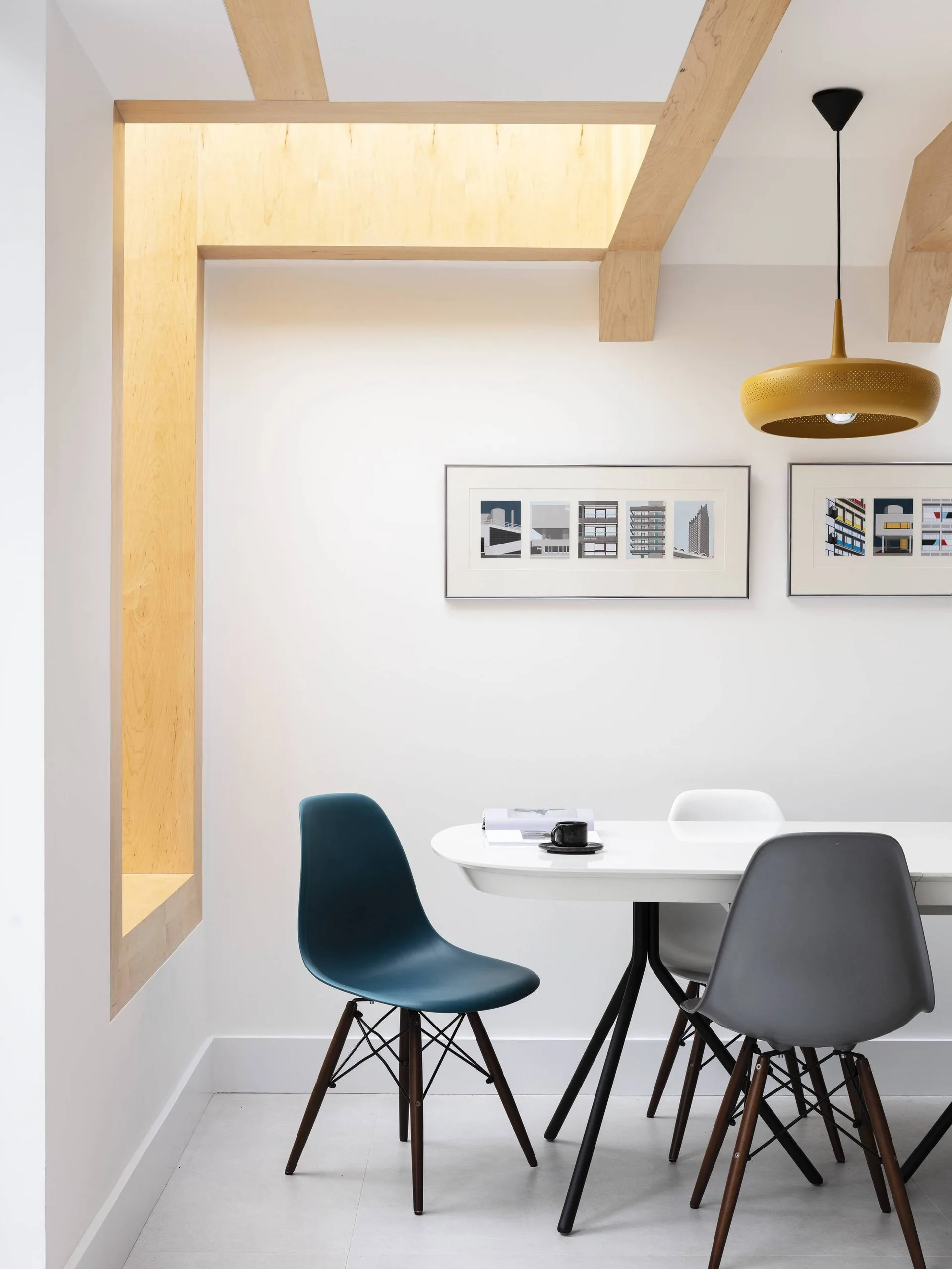 A modern dining area with a white oval table, colorful molded plastic chairs, exposed wood ceiling beams, and a yellow pendant light.