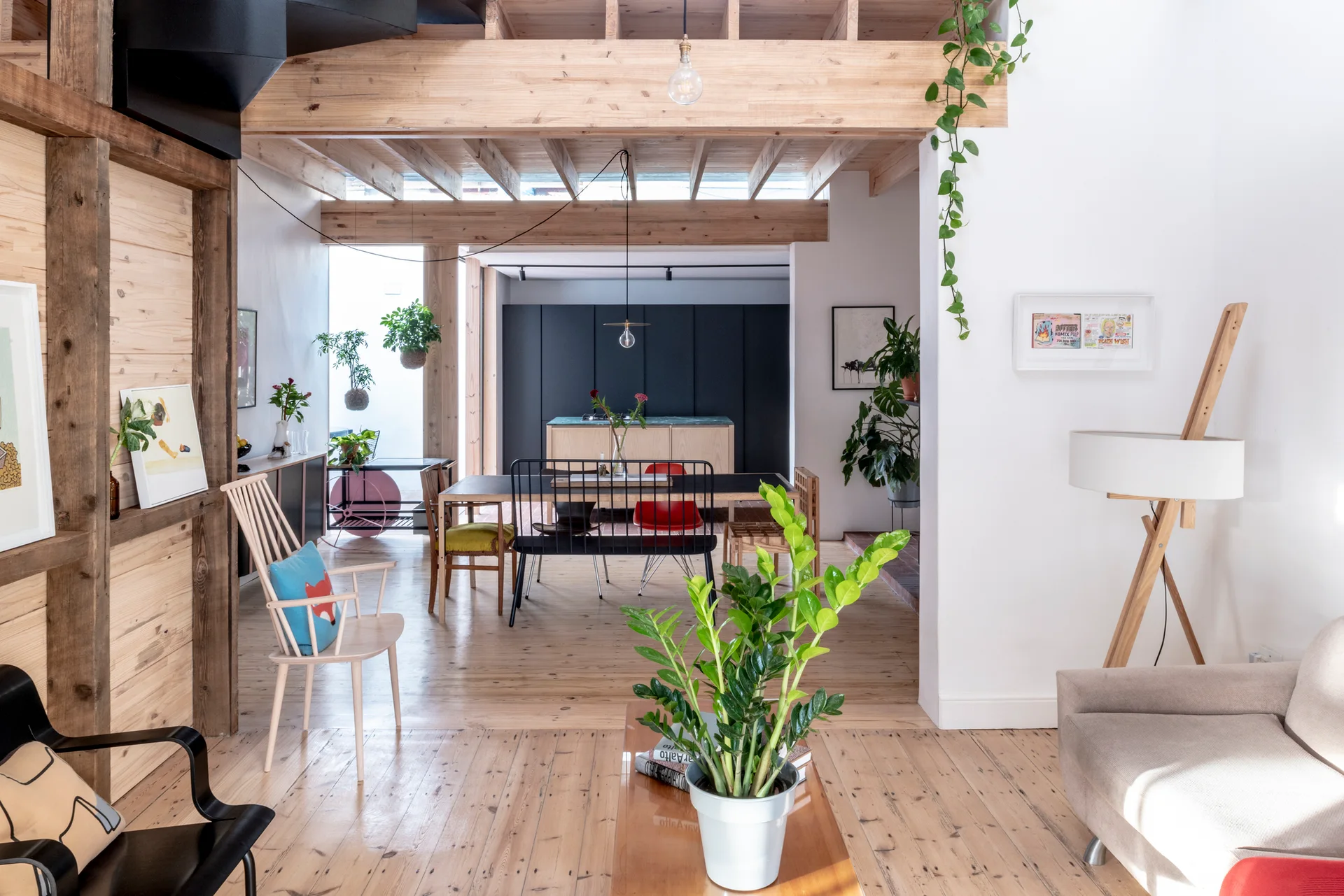 An open-plan dining and living space features exposed wooden ceiling beams, mismatched chairs around a long table, and light wood plank flooring.