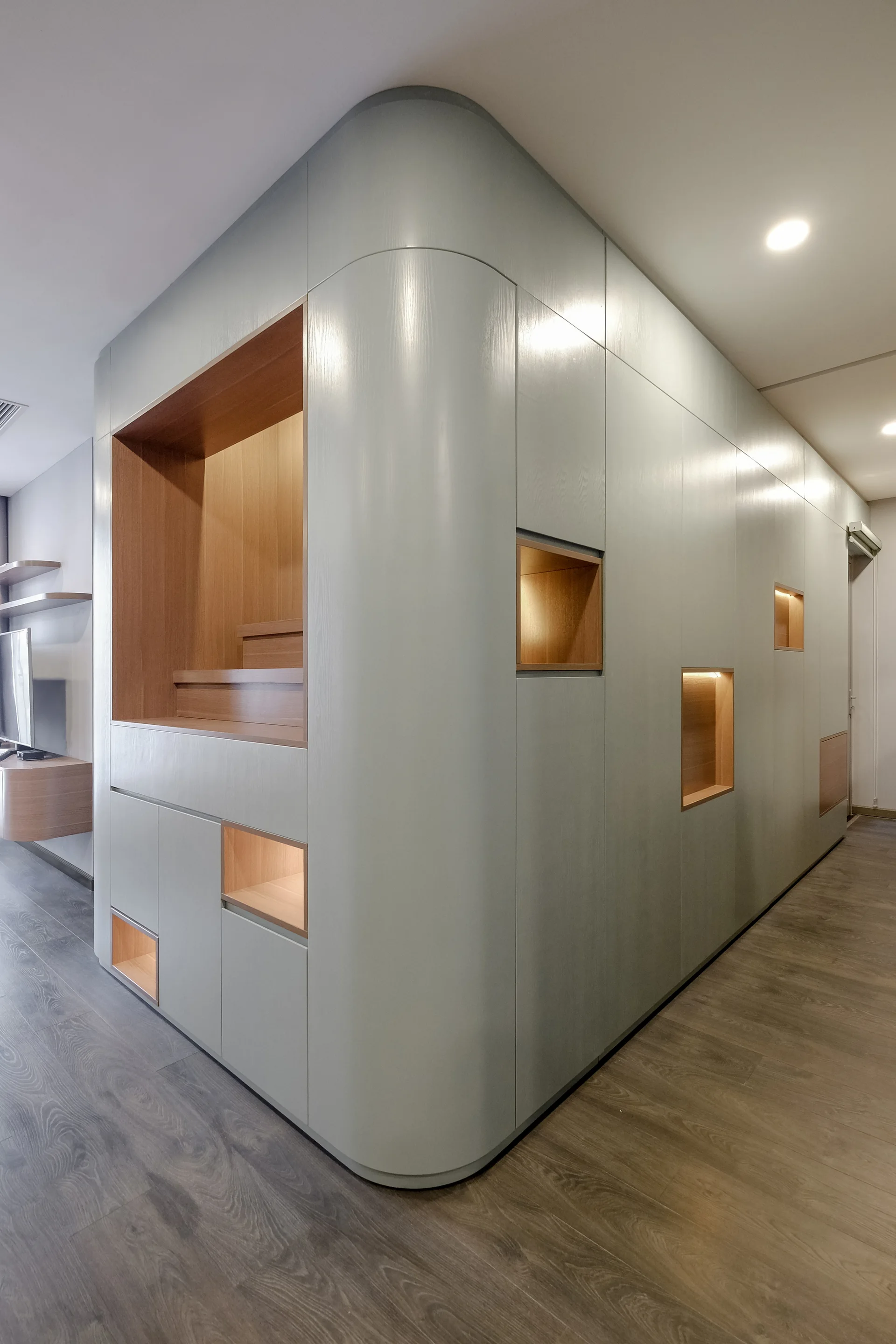 A floor-to-ceiling sage green cabinetry unit features a smooth curved corner and several wood-lined recessed niches illuminated with warm light.
