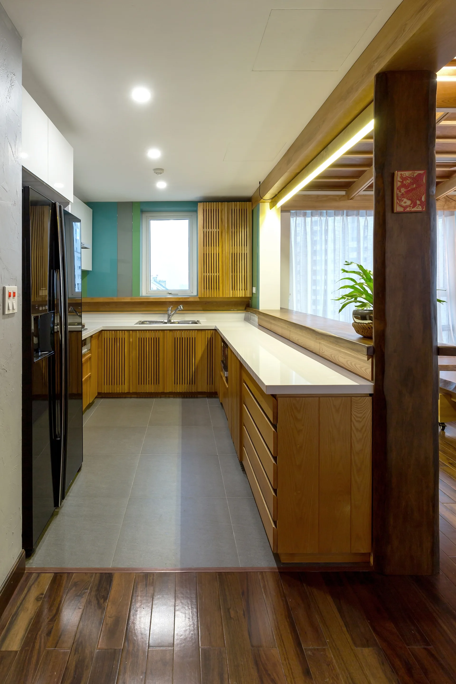 A modern kitchen with slatted light wood cabinets, white countertops, a black refrigerator, and a teal accent wall around the window.