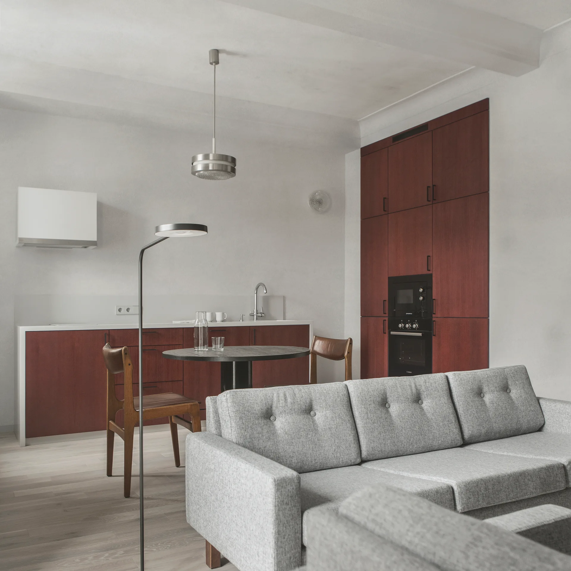 A modern open-plan room features cherry wood kitchen cabinets, a round dining table, and a grey tufted sofa in the foreground.