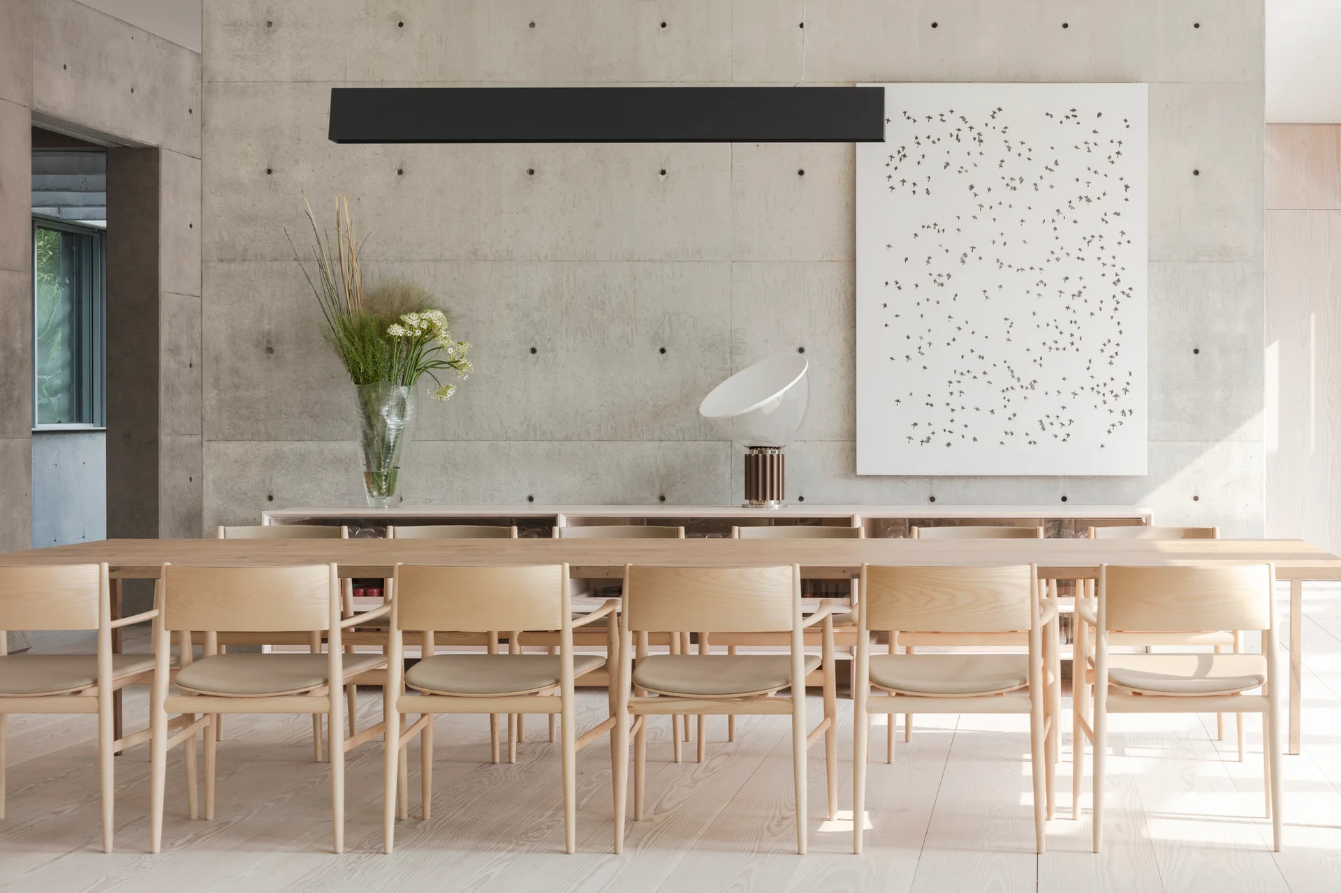 A long light wood dining table and matching chairs sit on wooden floors in front of an exposed concrete wall with a black linear pendant light.