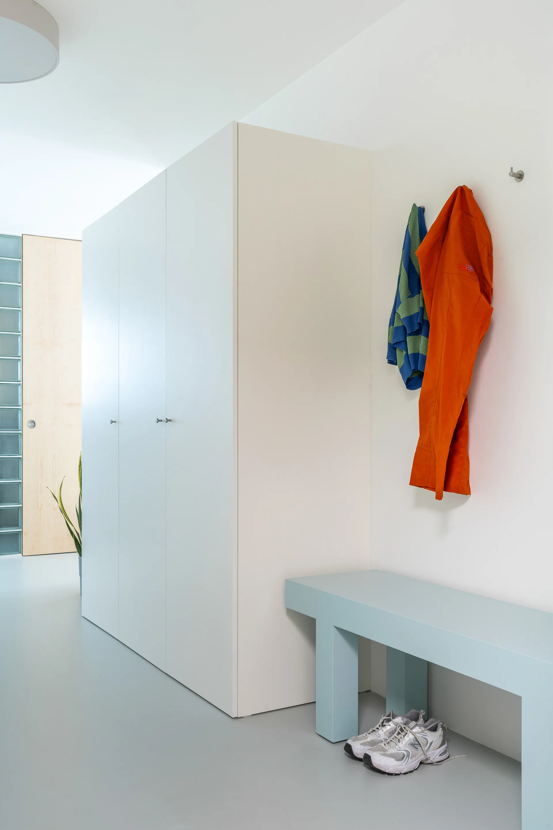 A minimalist entryway features a tall white wardrobe, a light blue bench with shoes underneath, and colorful jackets hanging on wall hooks.