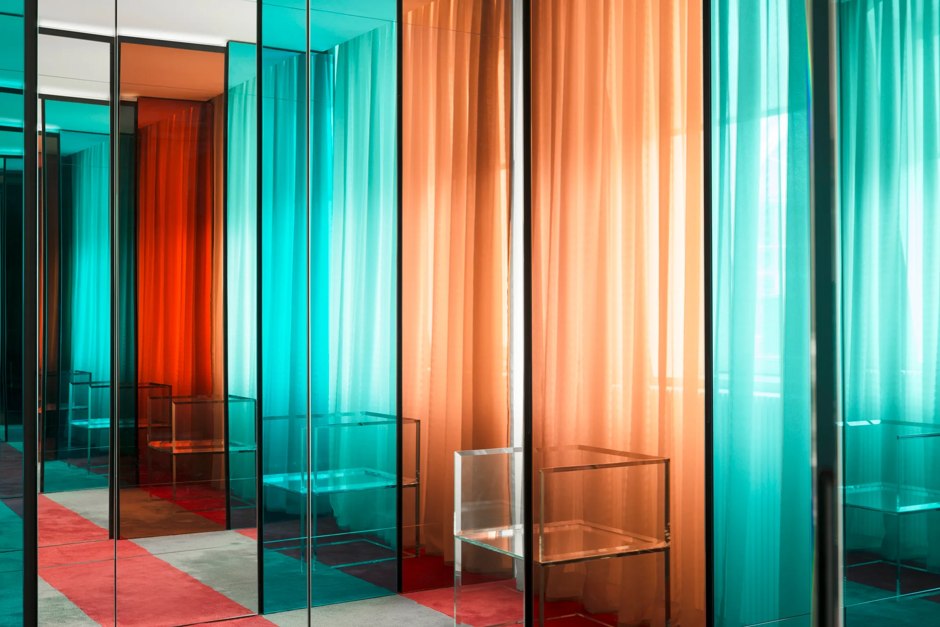 Tall teal and orange colored glass partitions reflect translucent sheer curtains and a clear acrylic box chair in a modern room.