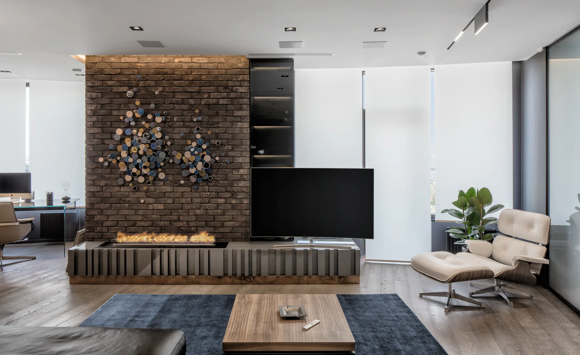 A modern living area with a dark brick wall featuring a built-in linear fireplace and TV, alongside a white lounge chair and blue rug.