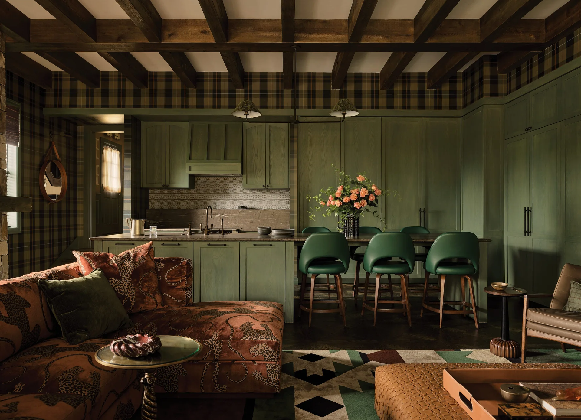 A dark-toned kitchen and living area featuring forest green cabinetry, plaid wallpaper, exposed wood ceiling beams, and a patterned sectional sofa.